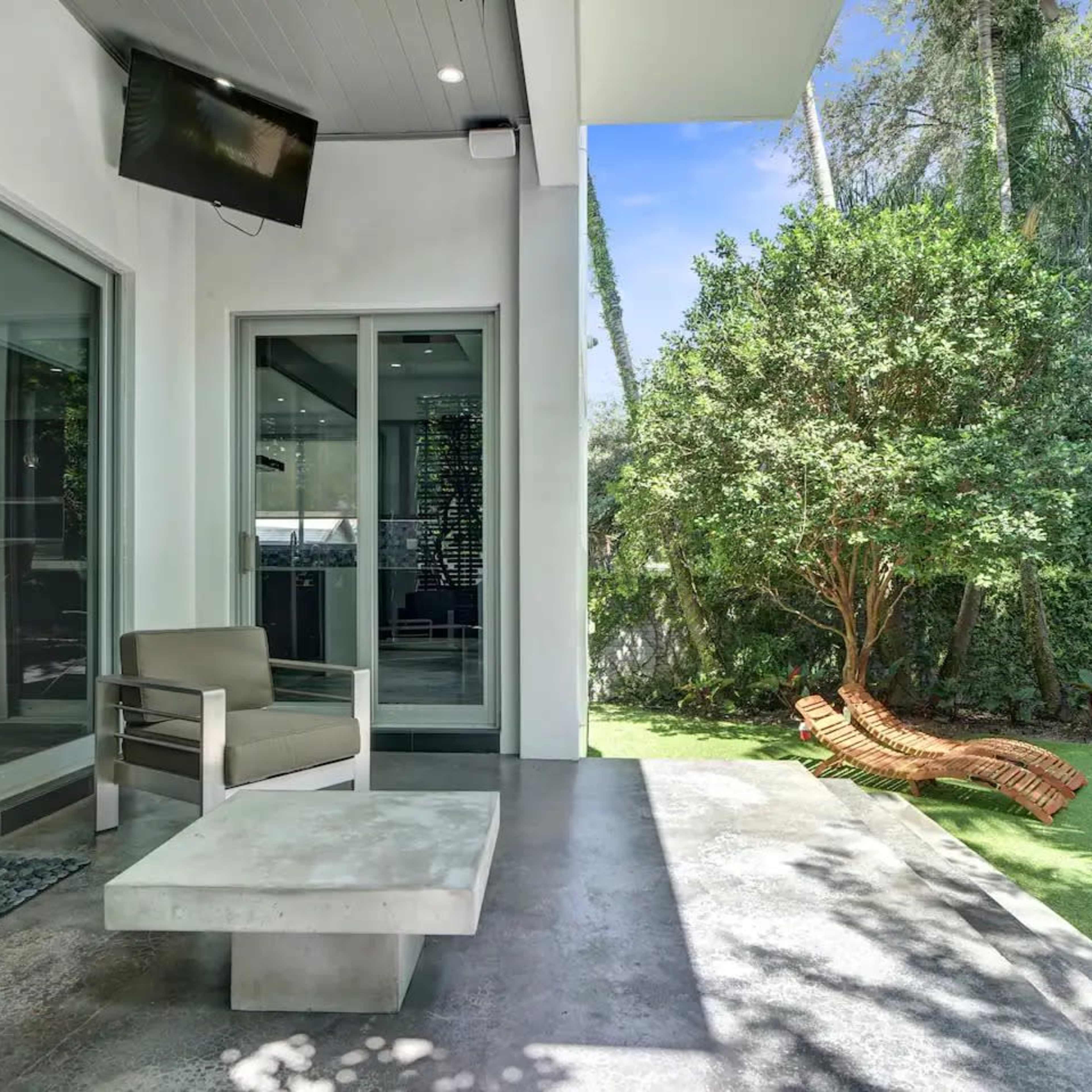 A patio area features modern seating, a coffee table, and a lush green garden with a wooden lounger and a barbecue grill.