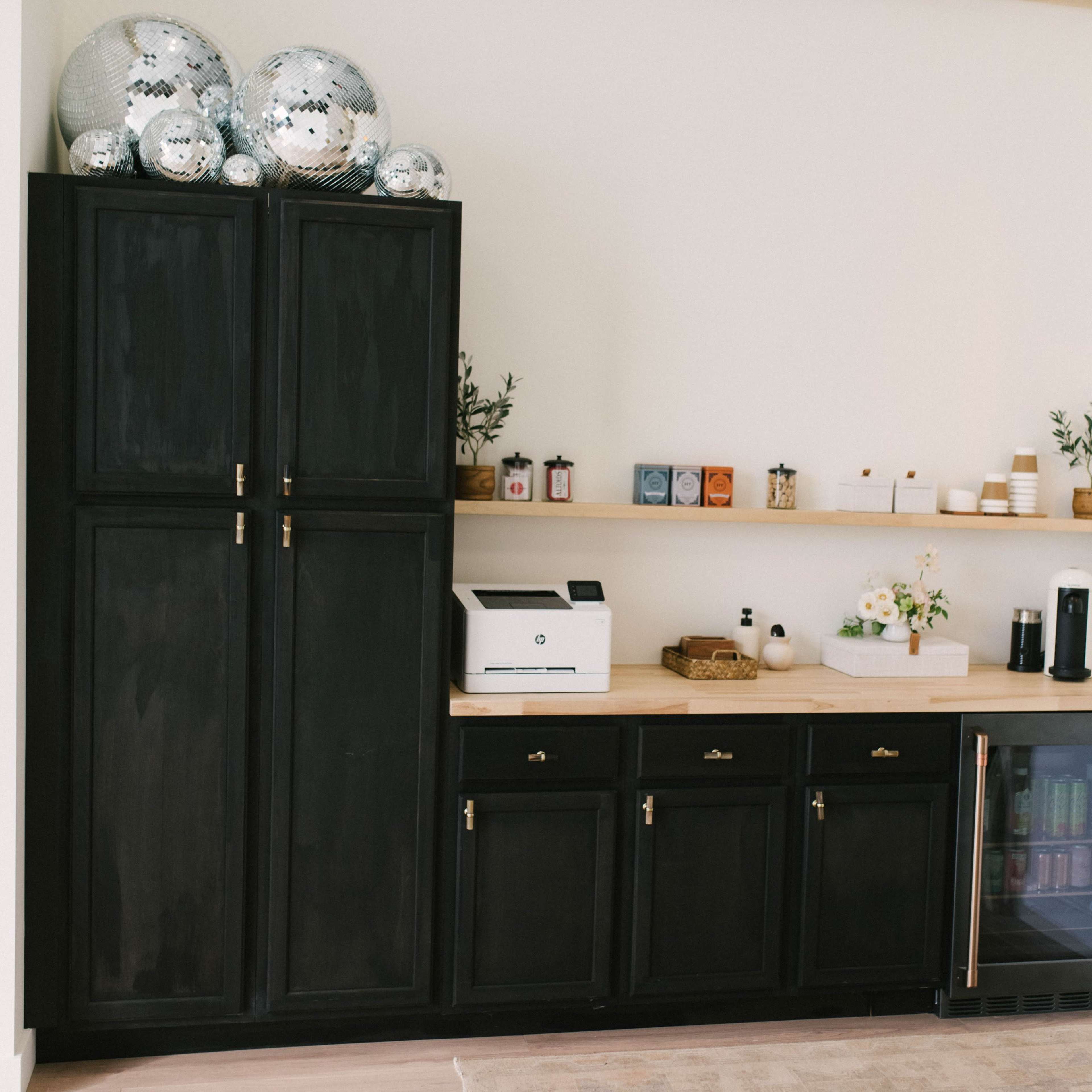 A modern kitchenette featuring black cabinetry, a countertop with decorative items, a printer, and a mini-fridge.