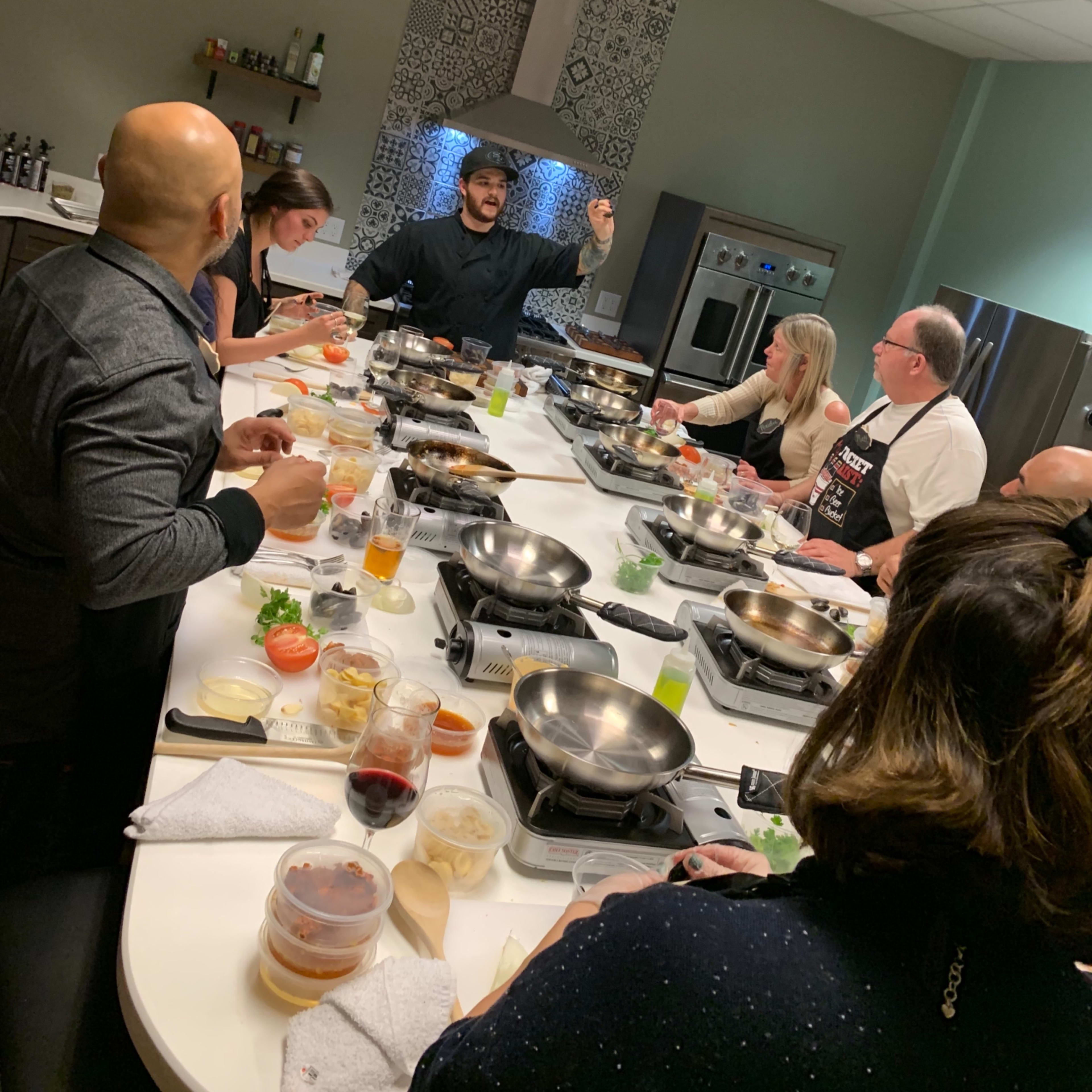 A diverse group of people gathers around a large, circular cooking station as a chef demonstrates a technique in a modern kitchen setting.