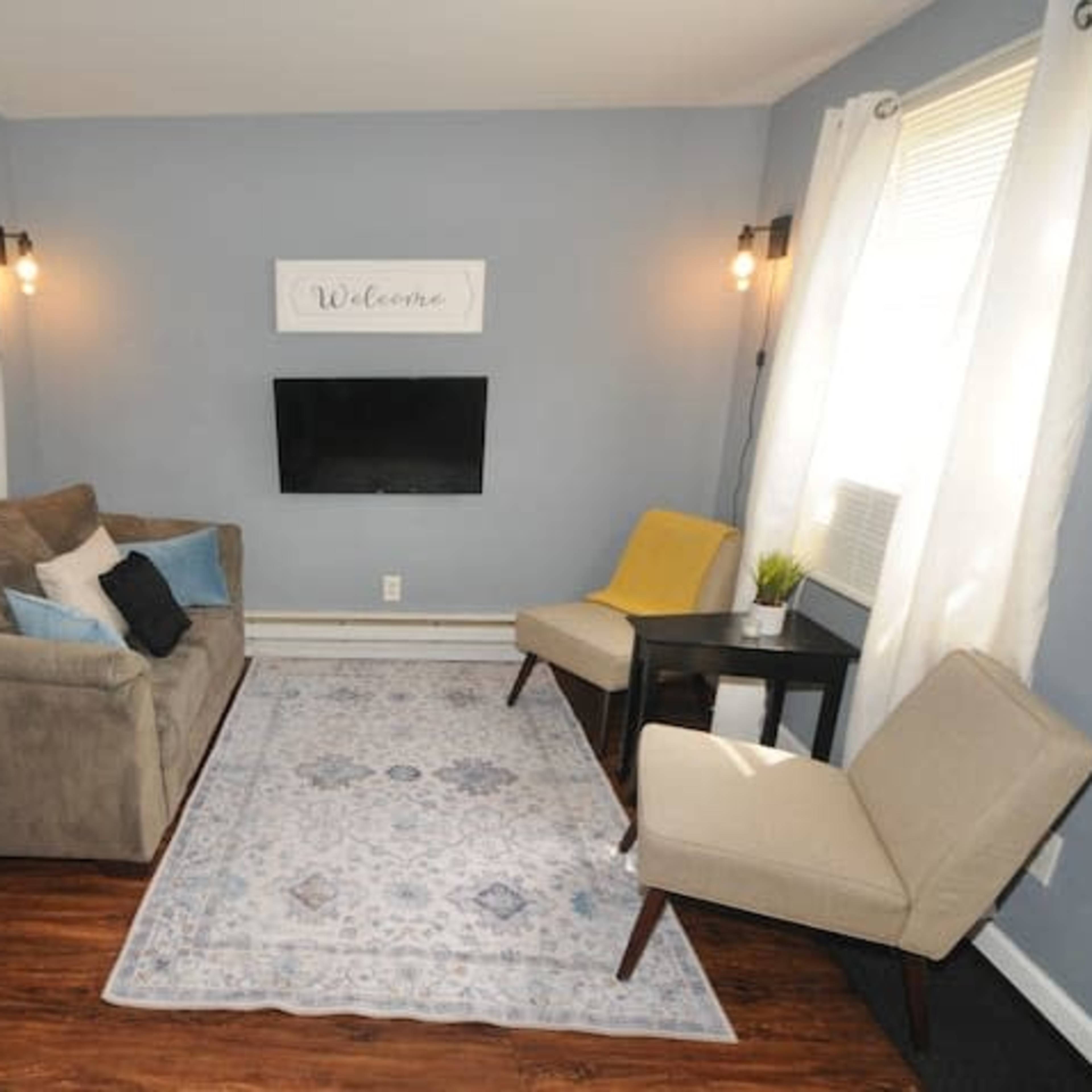 A small living room with a gray sofa, two chairs, a television mounted on the wall, and a decorative rug on a wooden floor.