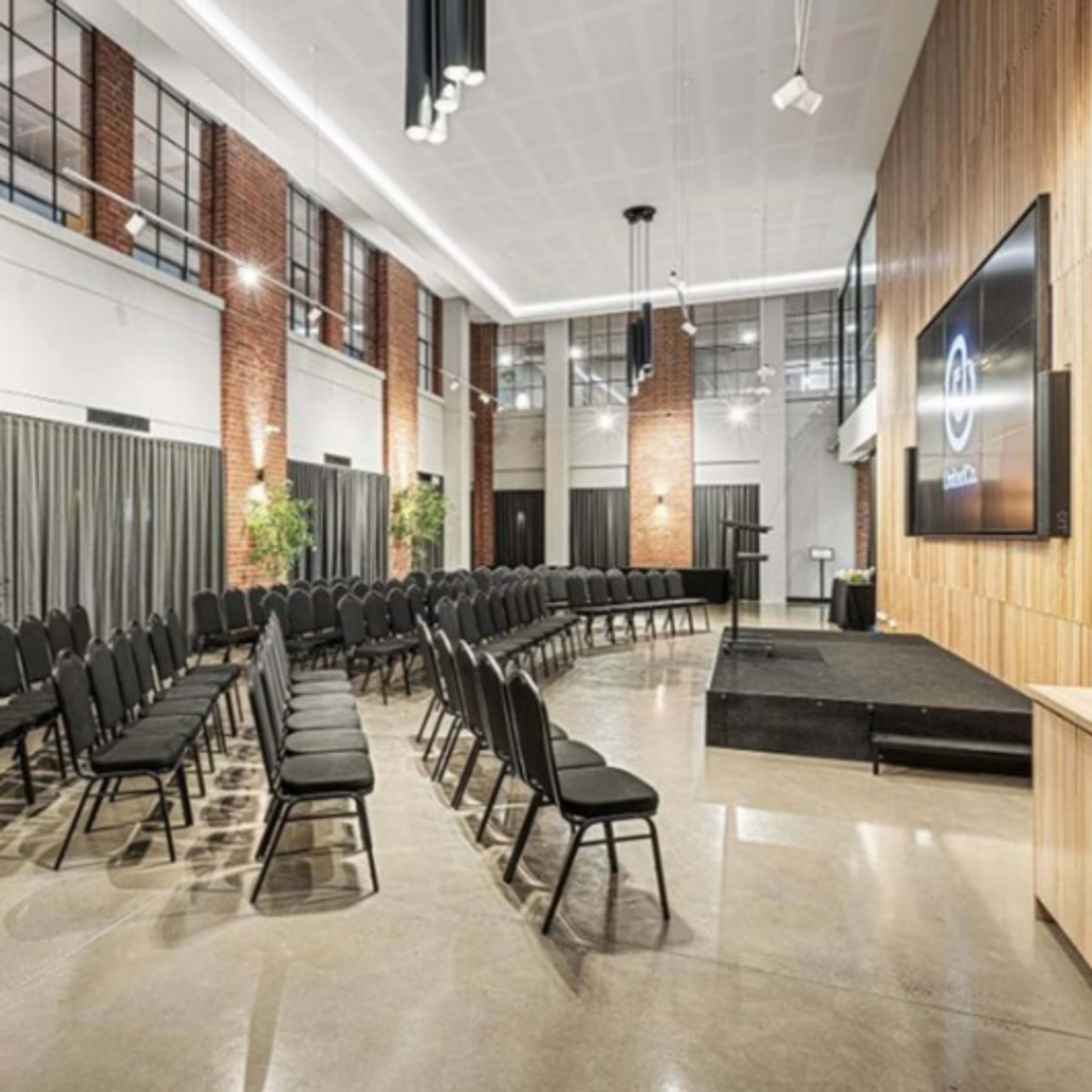 A modern event space features rows of black chairs facing a stage with wooden paneling, large windows, and potted plants.