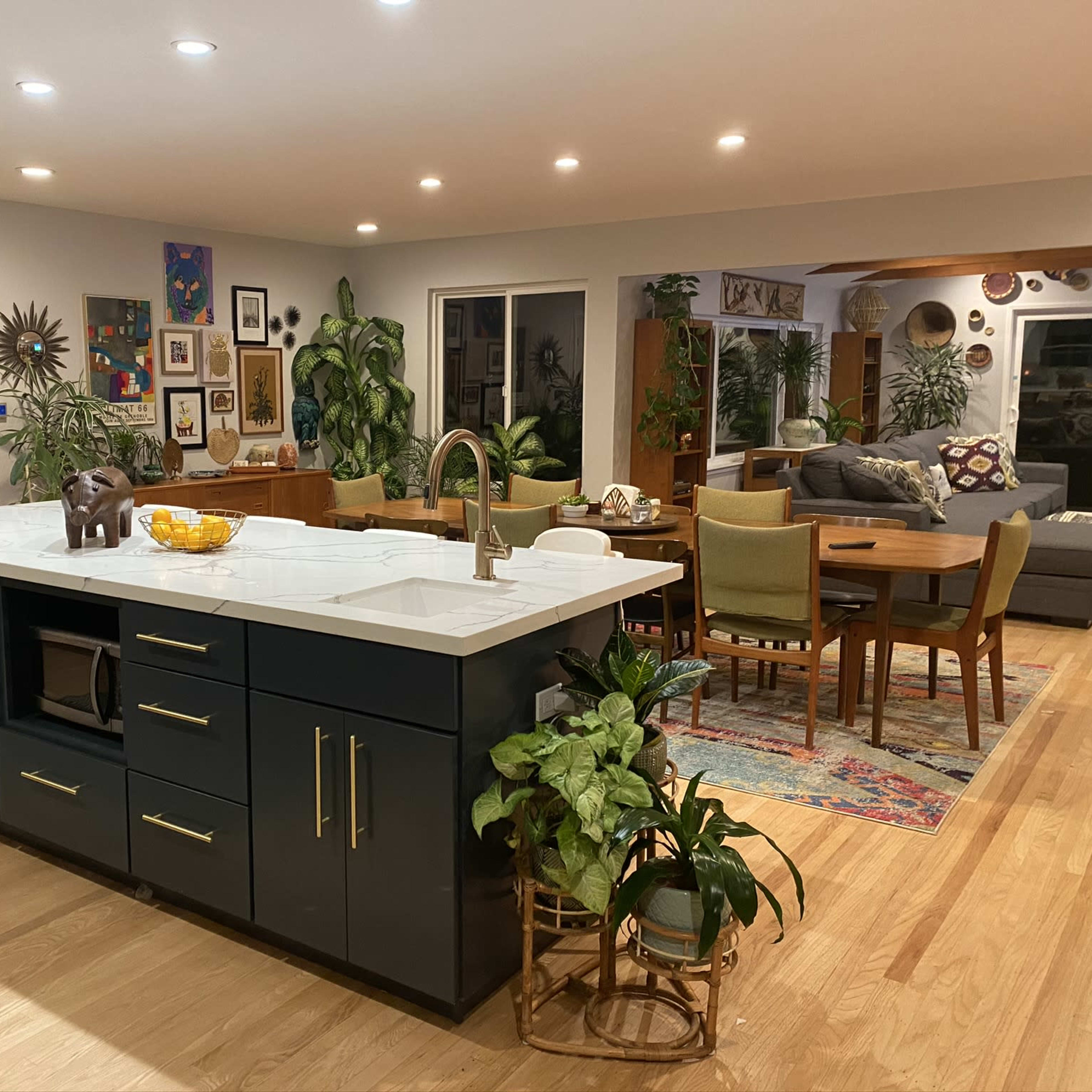 A modern kitchen with an island at the center, surrounded by dining and living areas featuring plants, artwork, and various furnishings.
