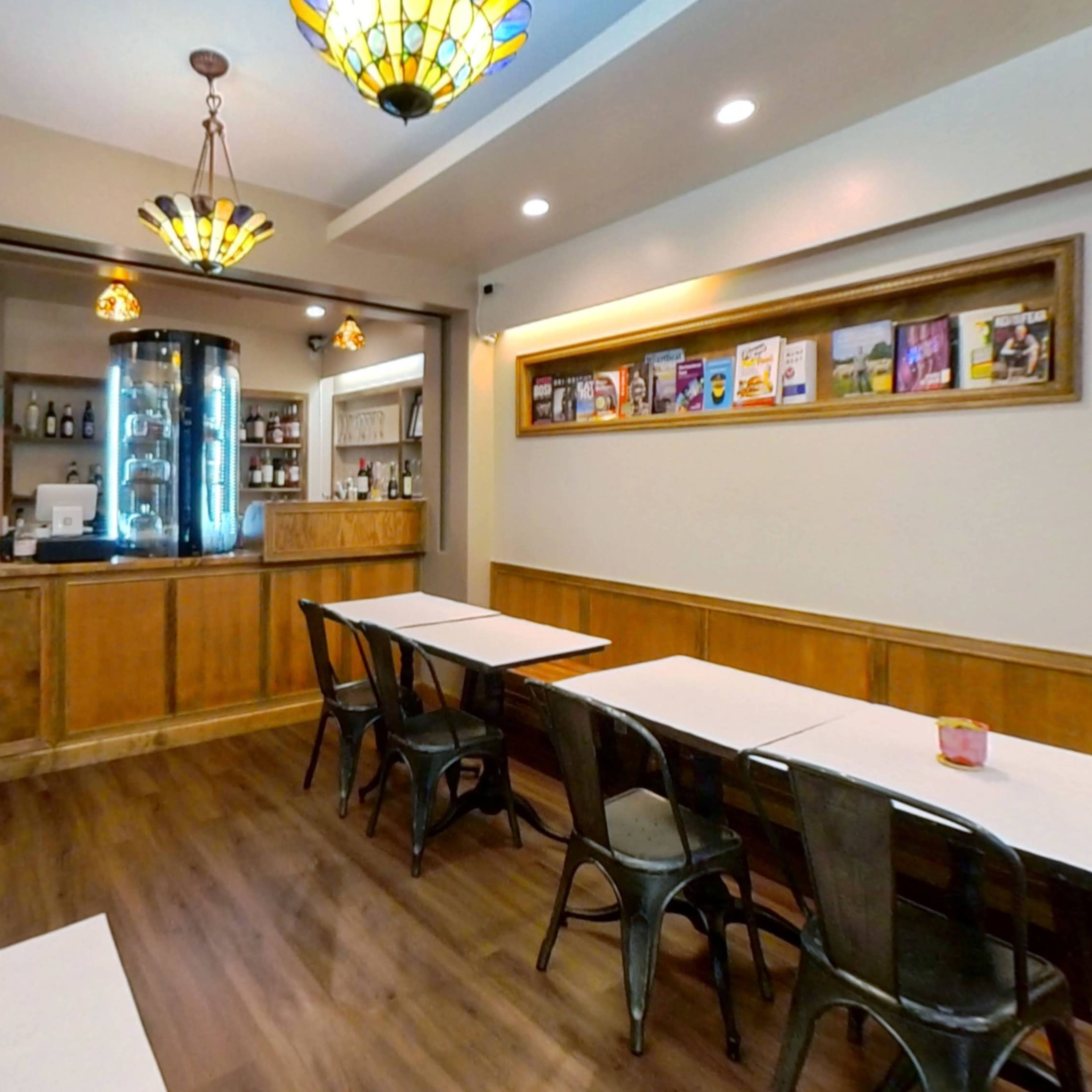 A dining area with several tables, metal chairs, and a wooden counter in a well-lit restaurant, featuring decorative light fixtures and a display shelf on the wall.