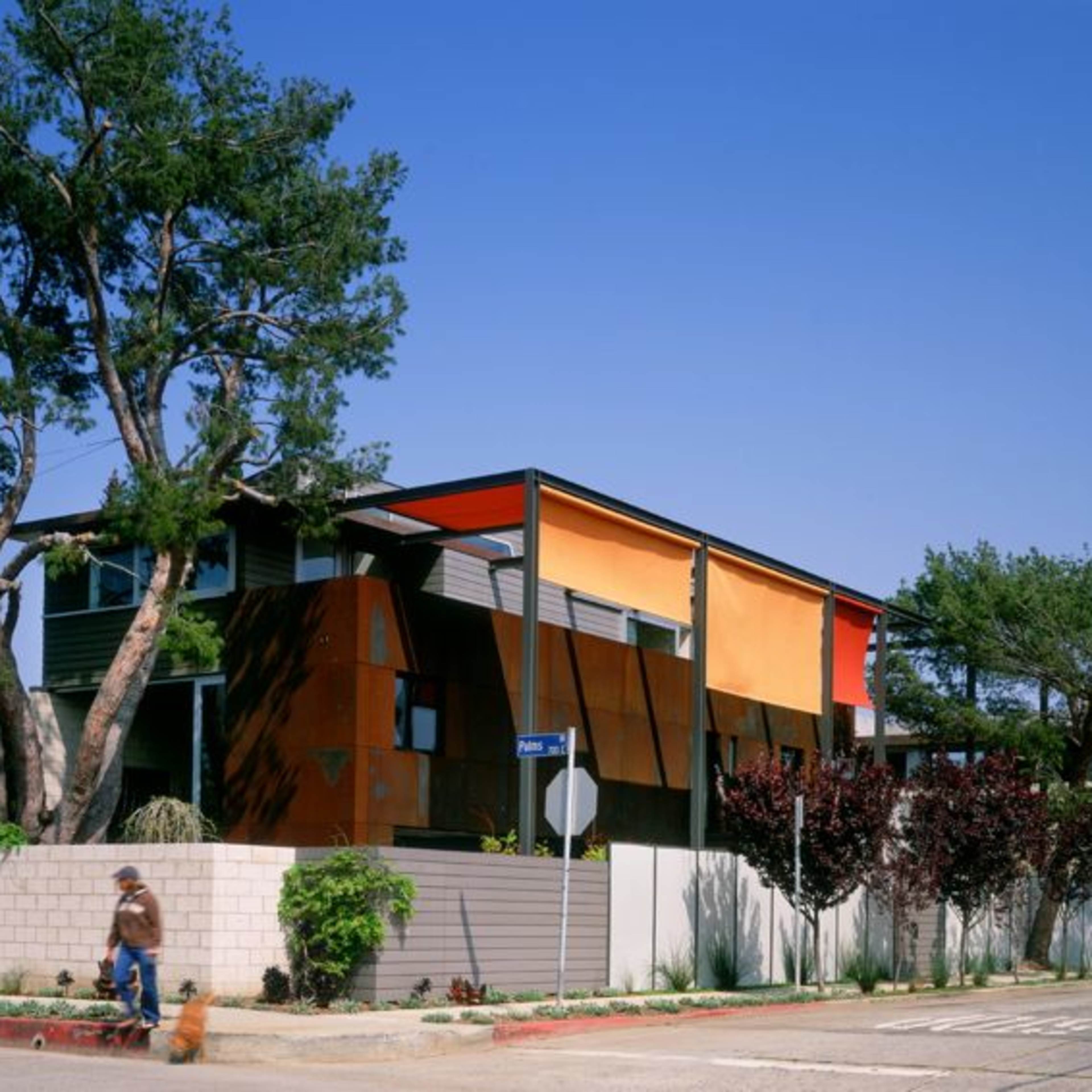 A modern house with orange and gray exterior panels is surrounded by trees and a low wall, with a person walking a dog along the sidewalk.