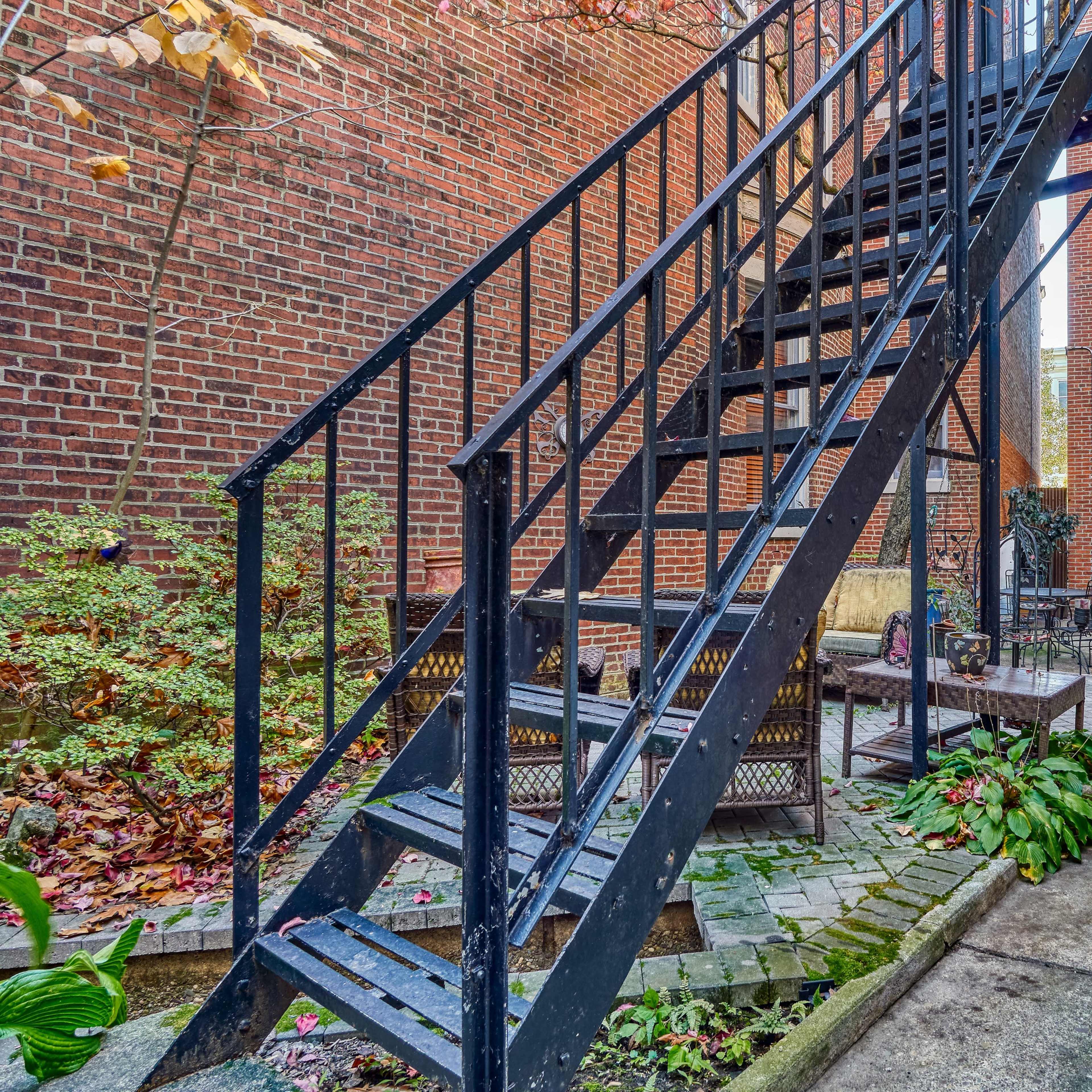 A courtyard with a metal stairway leading to an upper level, flanked by brick walls and outdoor furniture among greenery.