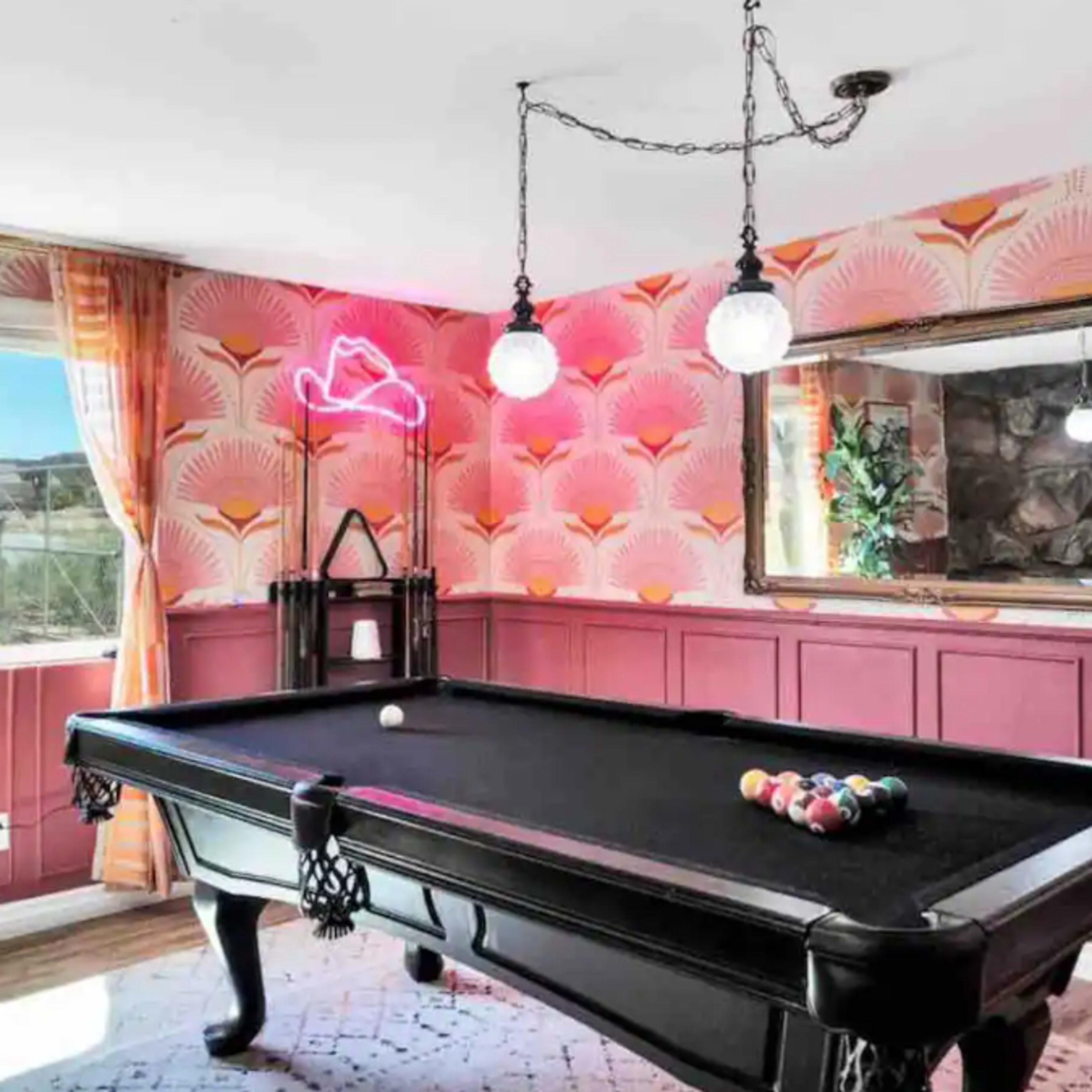 A billiard room features a black pool table with a set of colored balls arranged, surrounded by patterned wallpaper and modern lighting fixtures.