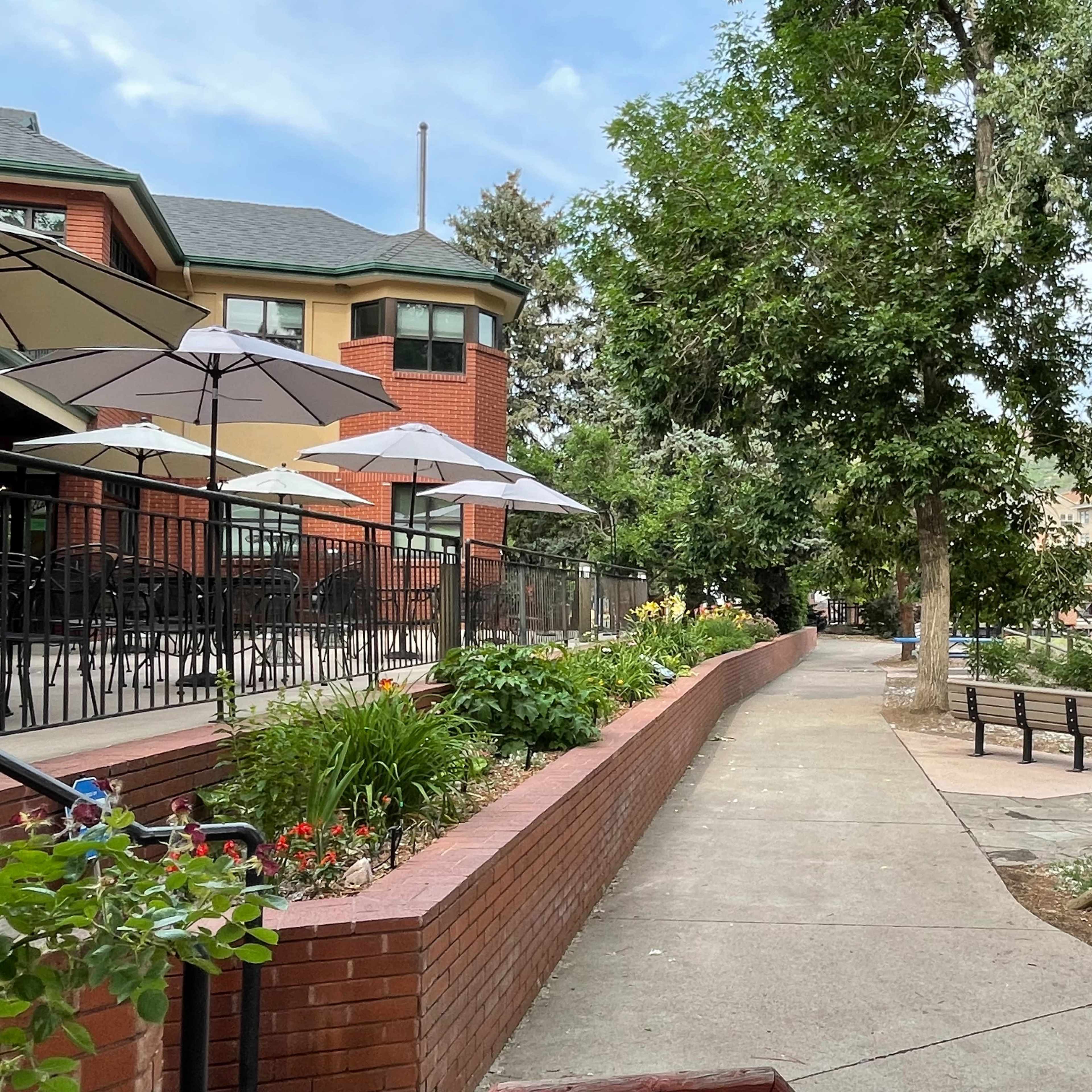 A paved walkway with landscaped flower beds runs alongside a building with outdoor seating under umbrellas.