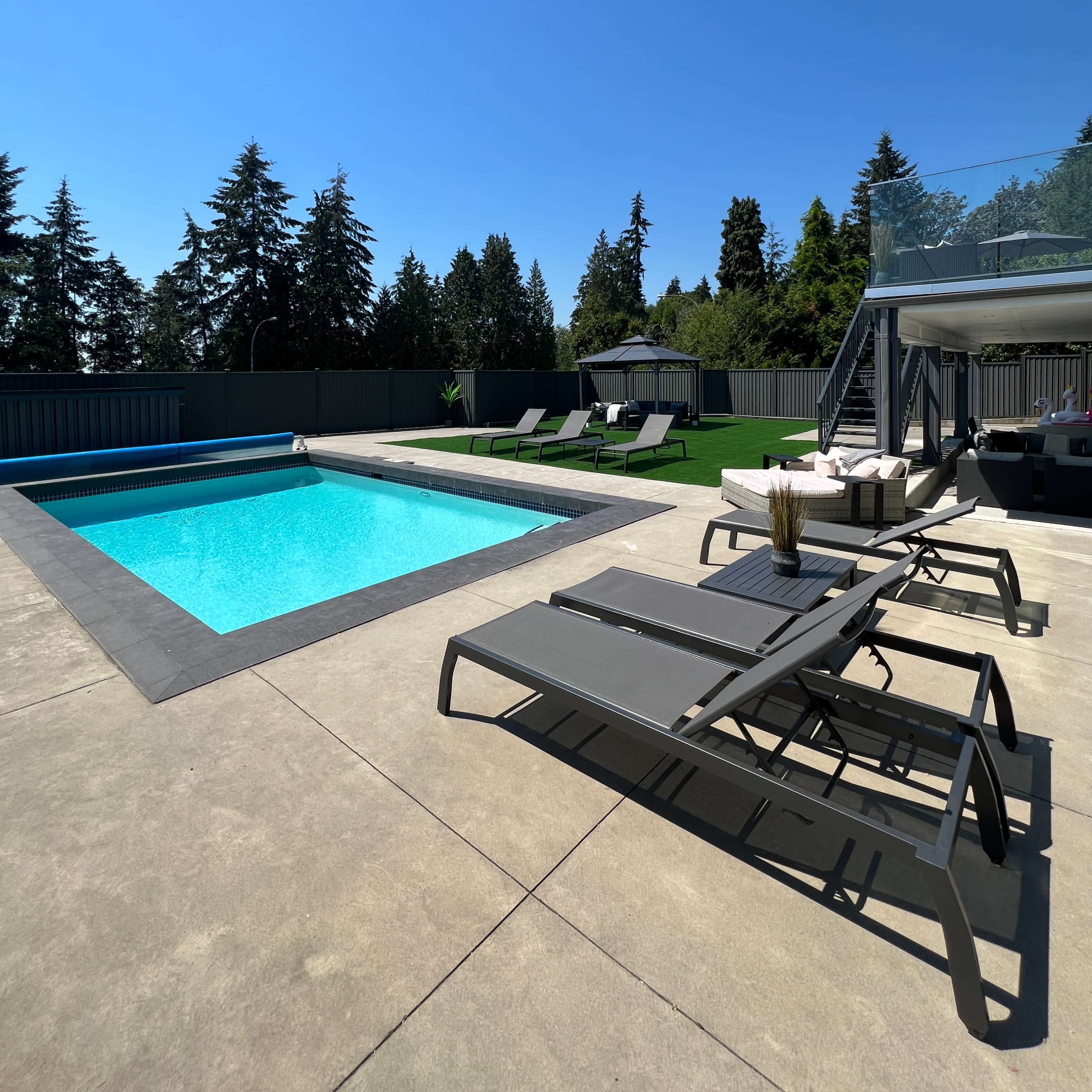 A rectangular swimming pool is surrounded by lounge chairs and a patio area, with trees and a shed in the background under a clear blue sky.