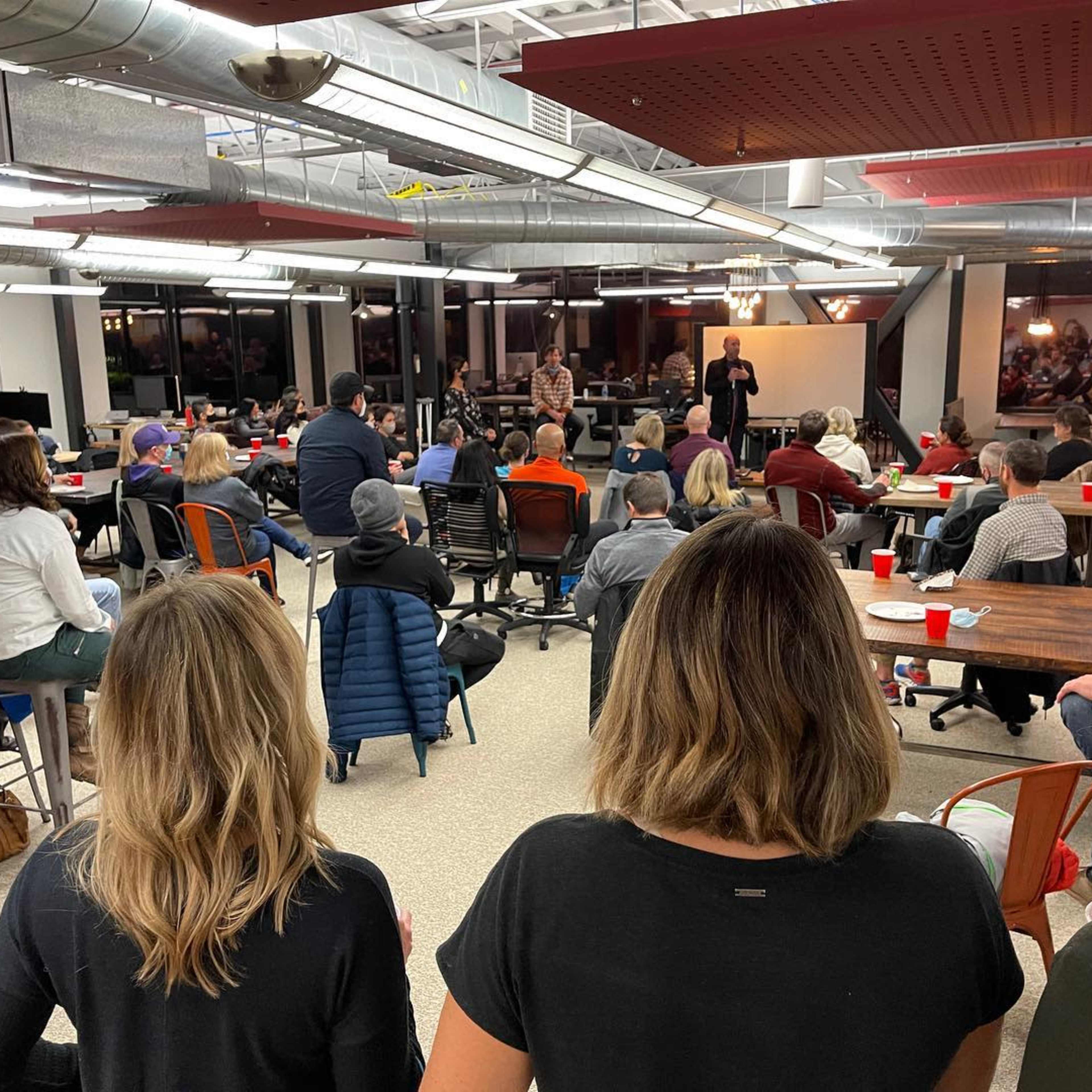 A group of people is seated in a large, open room, facing a speaker at the front, with tables and chairs arranged for an event.