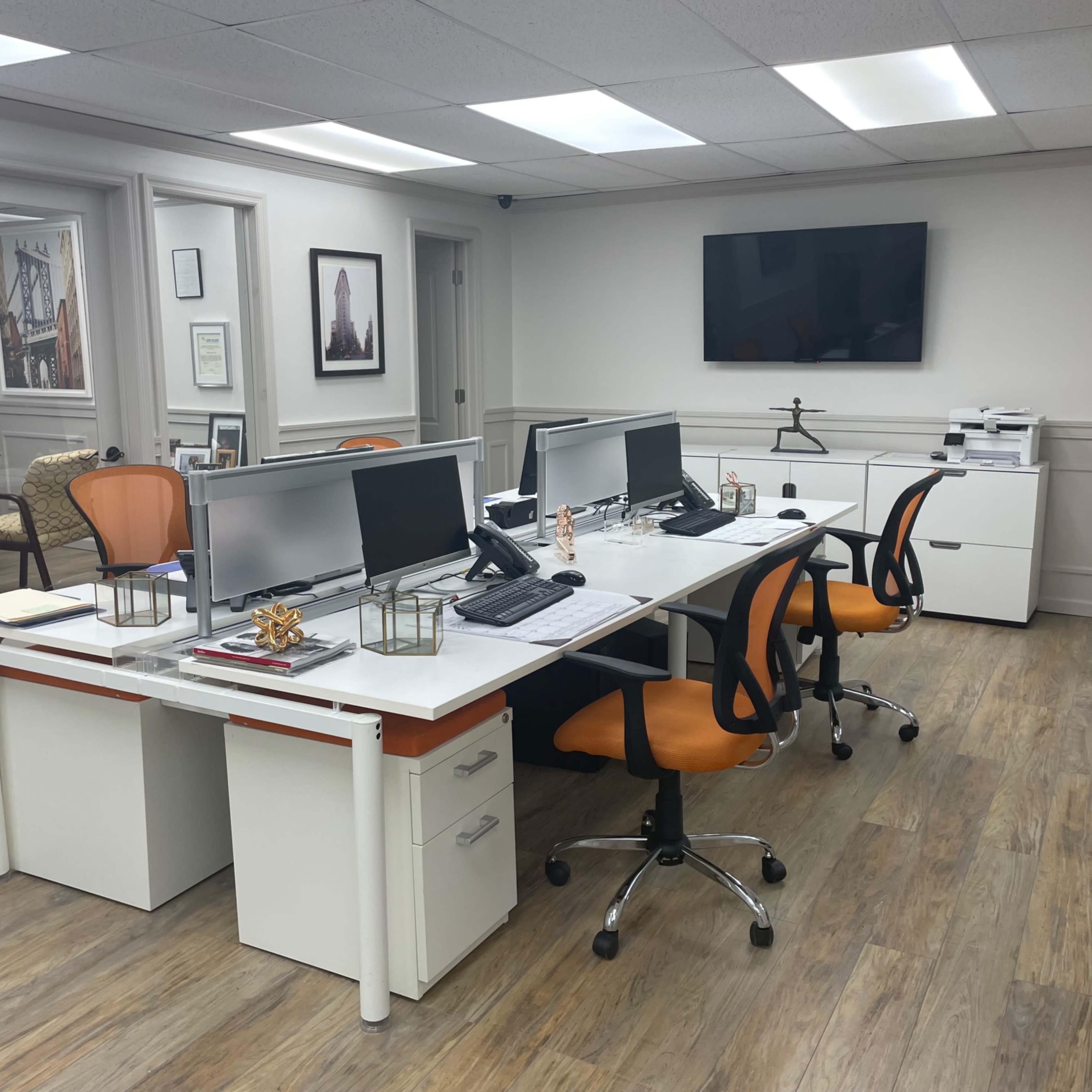 A modern office space with two white desks, each equipped with dual computer monitors, surrounded by orange office chairs, and a printer station in the background.