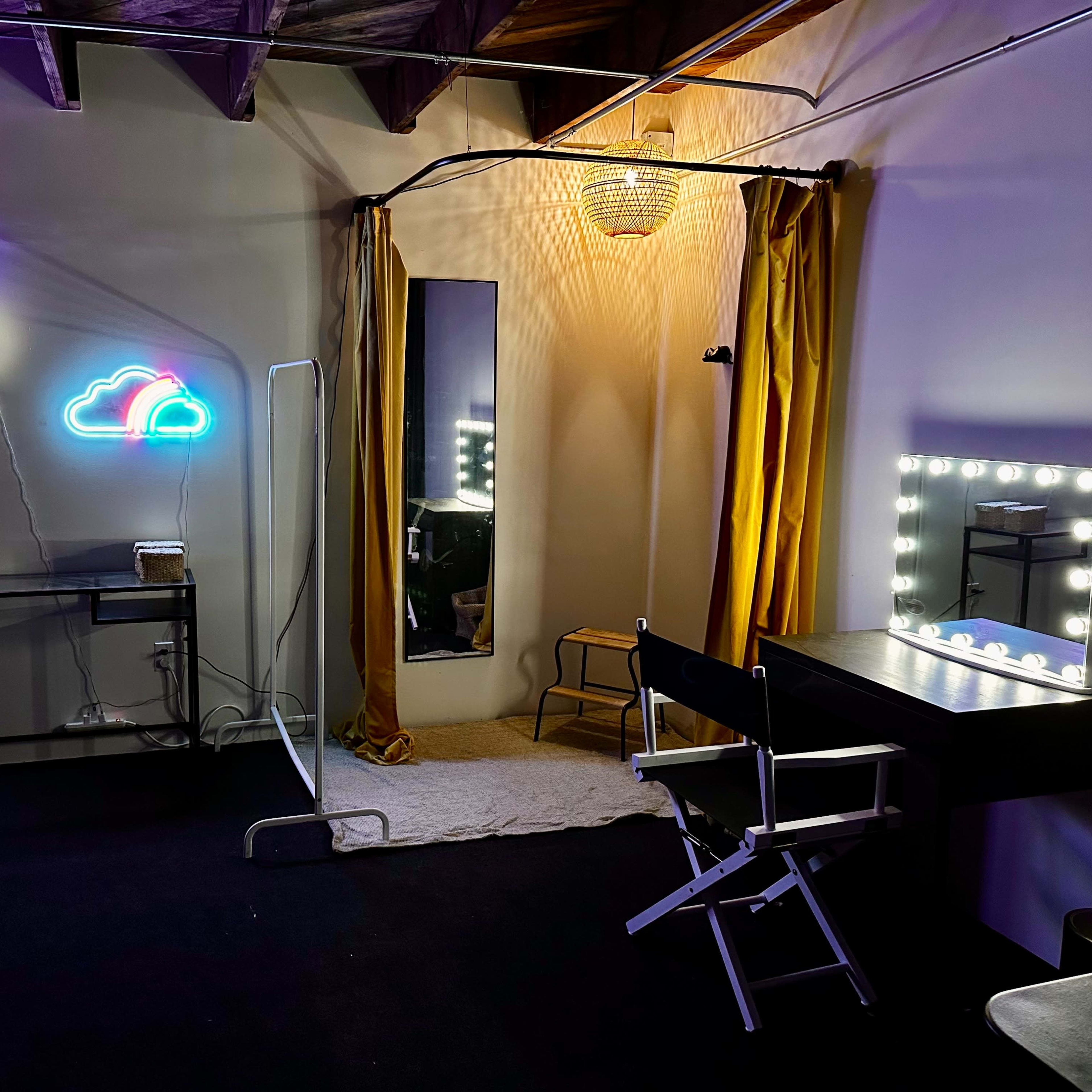 A dimly lit room featuring a makeup vanity with bright lights, a mirror, a curtain, and neon cloud decorations on the walls.