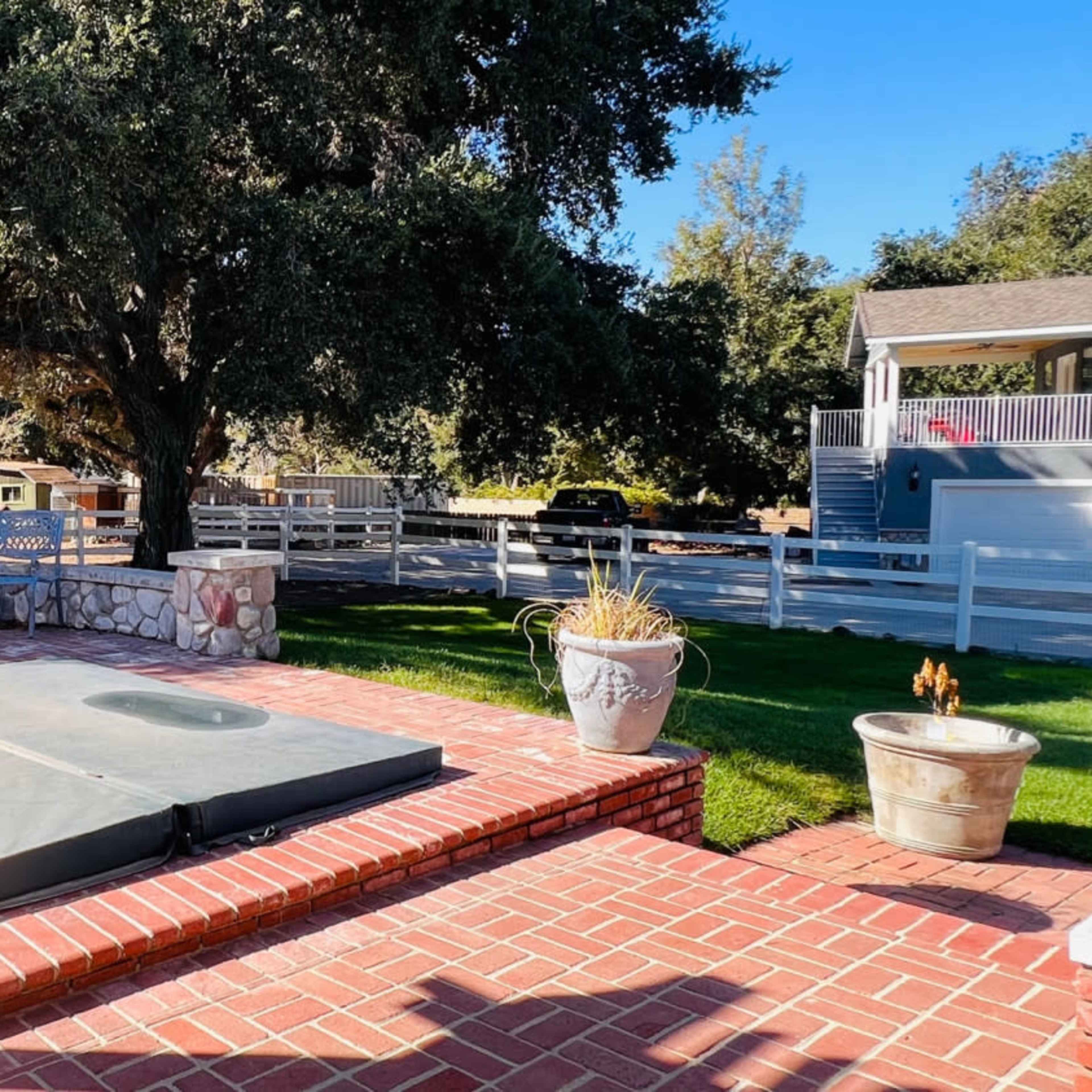 A patio area features a covered hot tub surrounded by brick paving, stone seating, and lush greenery under a large tree.
