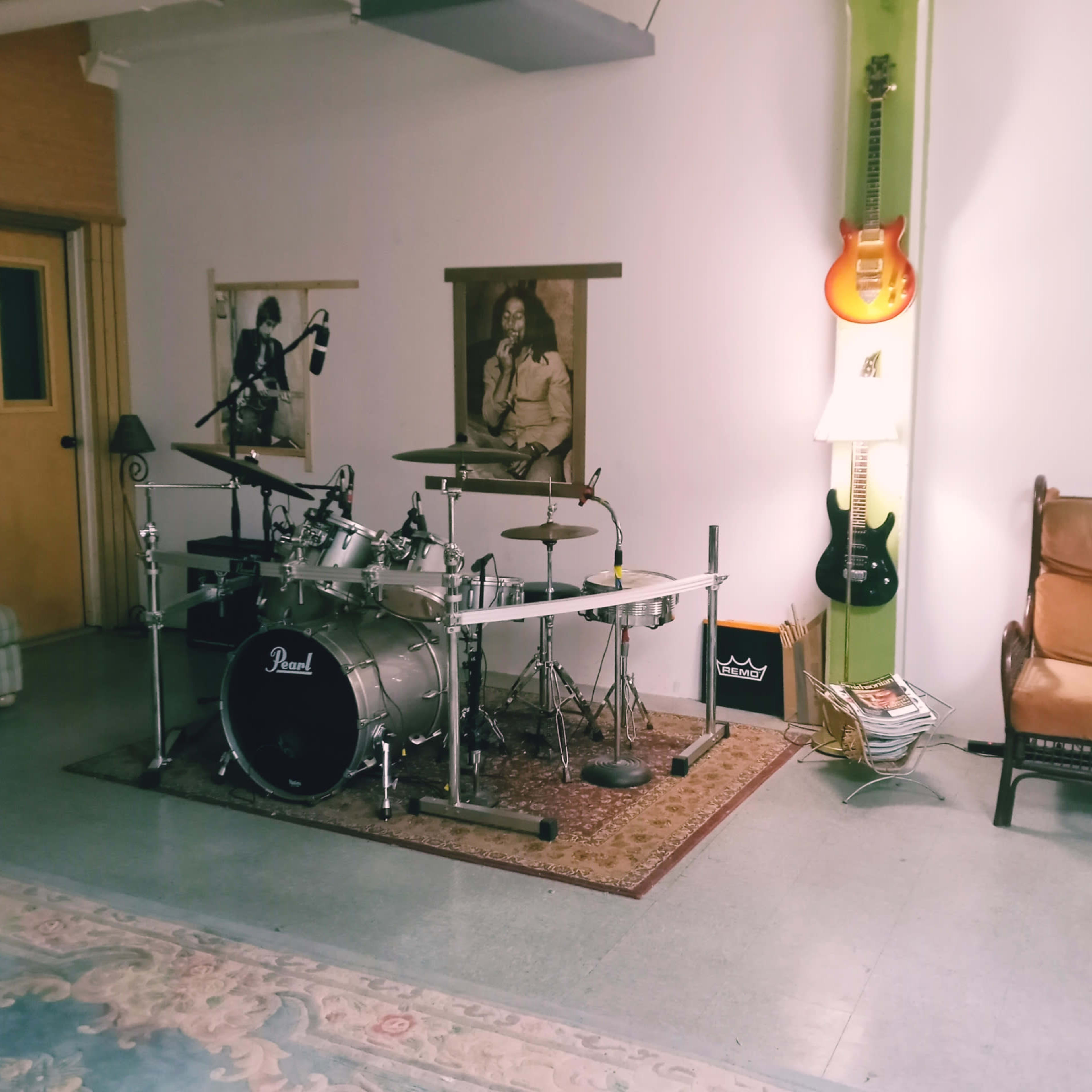 A music practice room featuring a drum set, electric guitars mounted on the wall, and a chair beside a lamp.