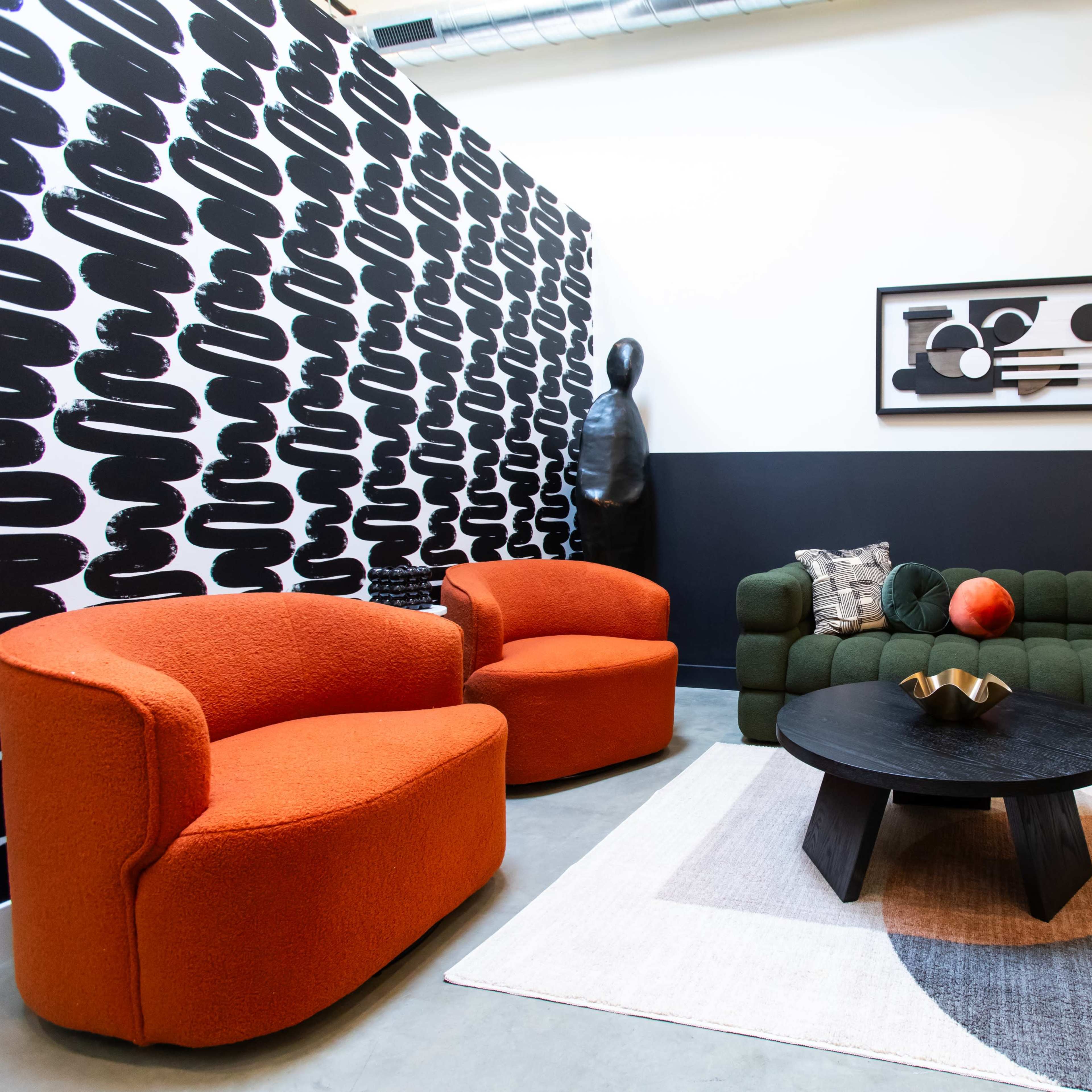 A modern seating area features two orange armchairs and a green sofa arranged around a round black coffee table on a patterned rug, against a black and white patterned wall.