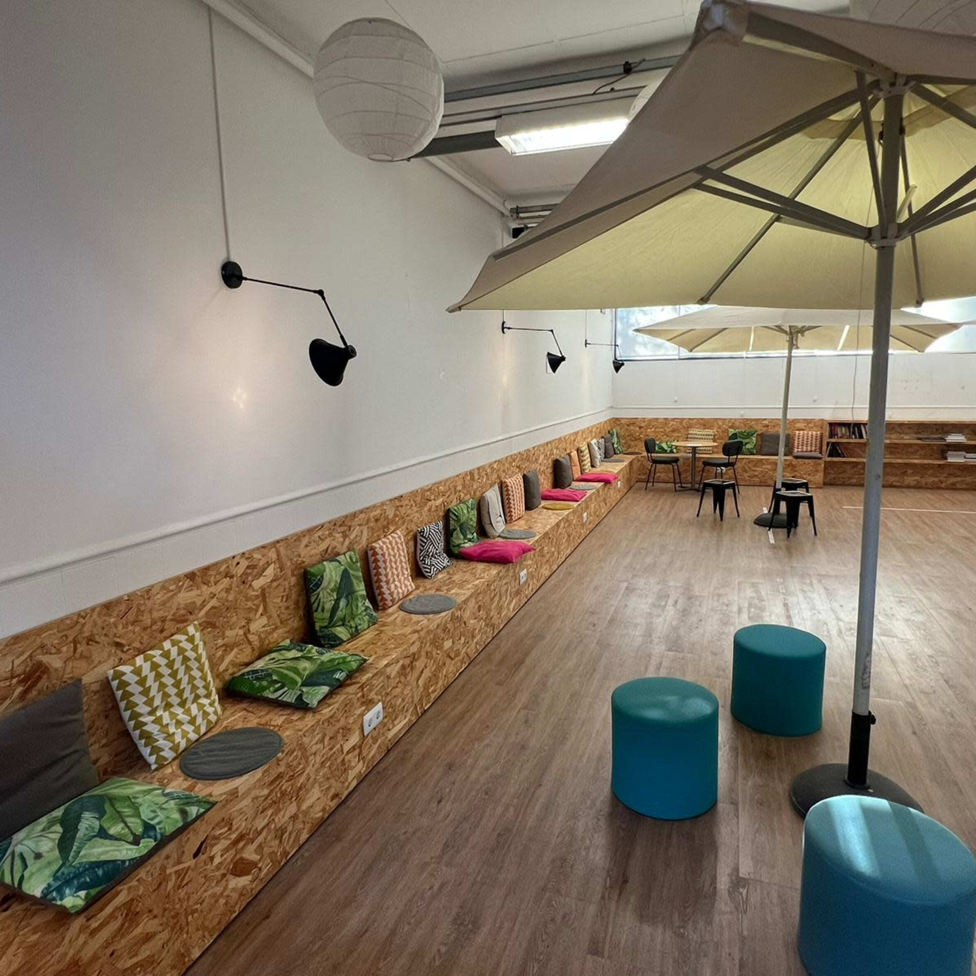 A spacious room with a long wooden bench featuring colorful cushions and several round stools, illuminated by wall-mounted lamps and large umbrellas.