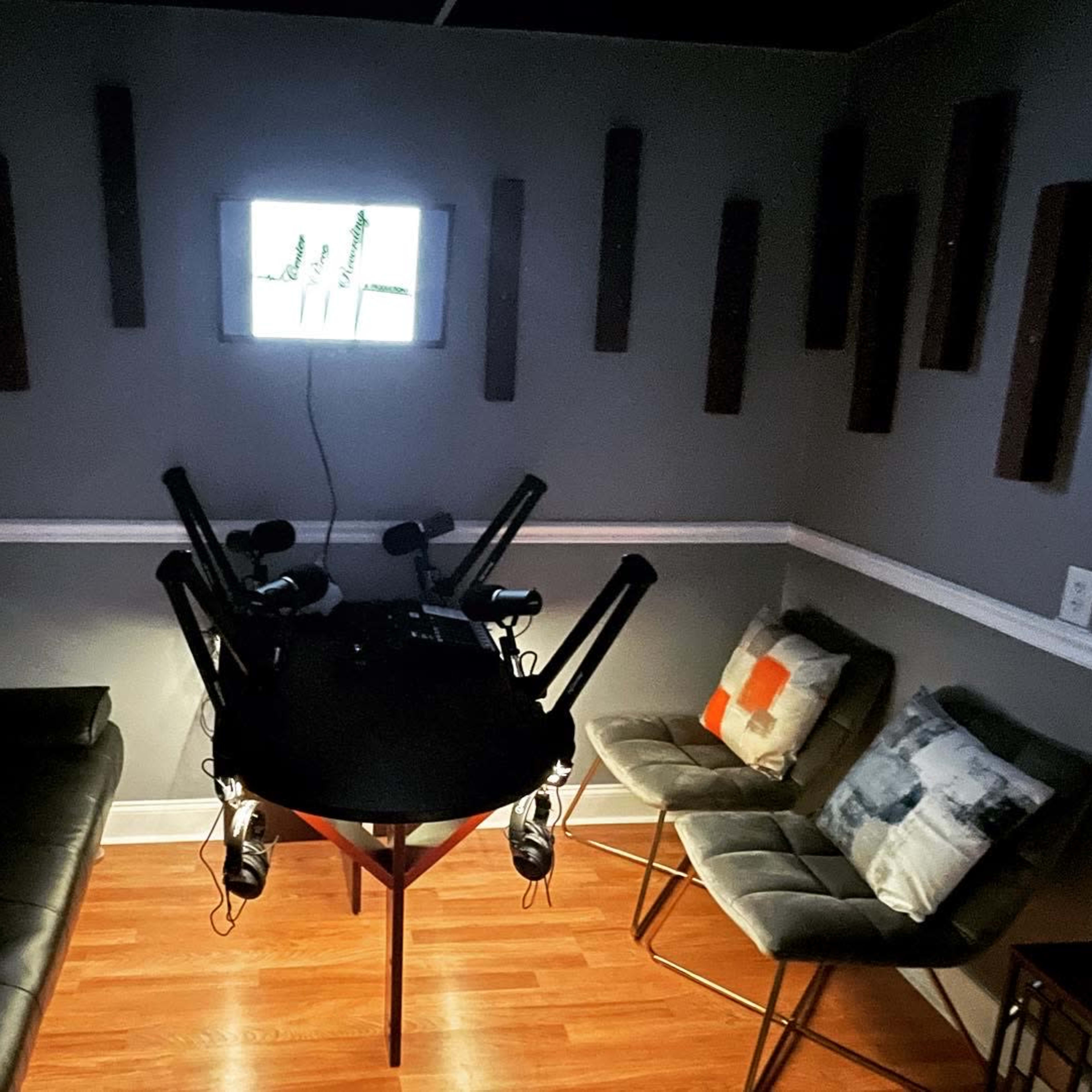 A small room with a black sofa, a round table equipped with microphones, and two chairs, surrounded by wall panels and a window displaying a bright scene.