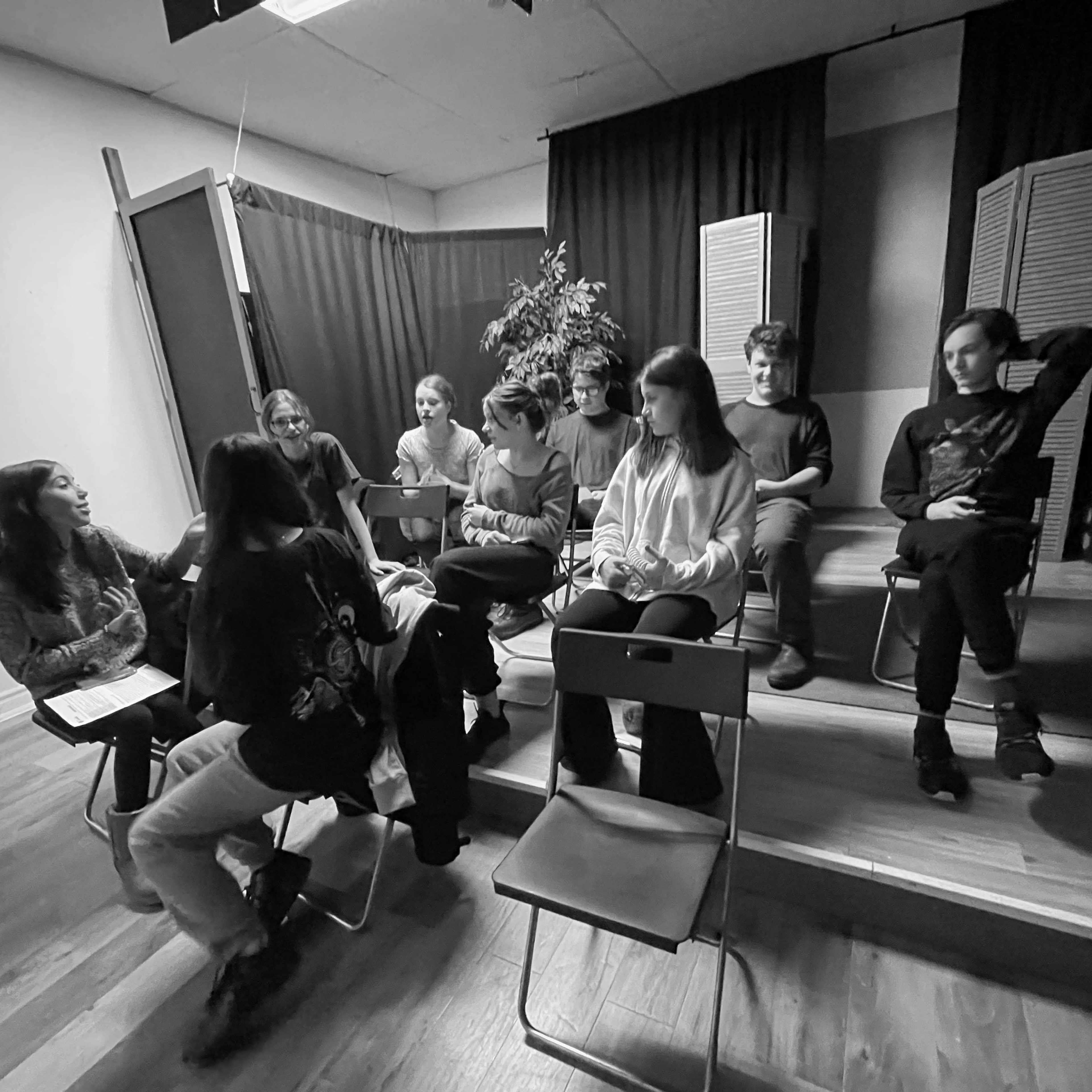 A group of eight people sits in chairs arranged in a semi-circle, with one person speaking at the front of a small, indoor stage area.