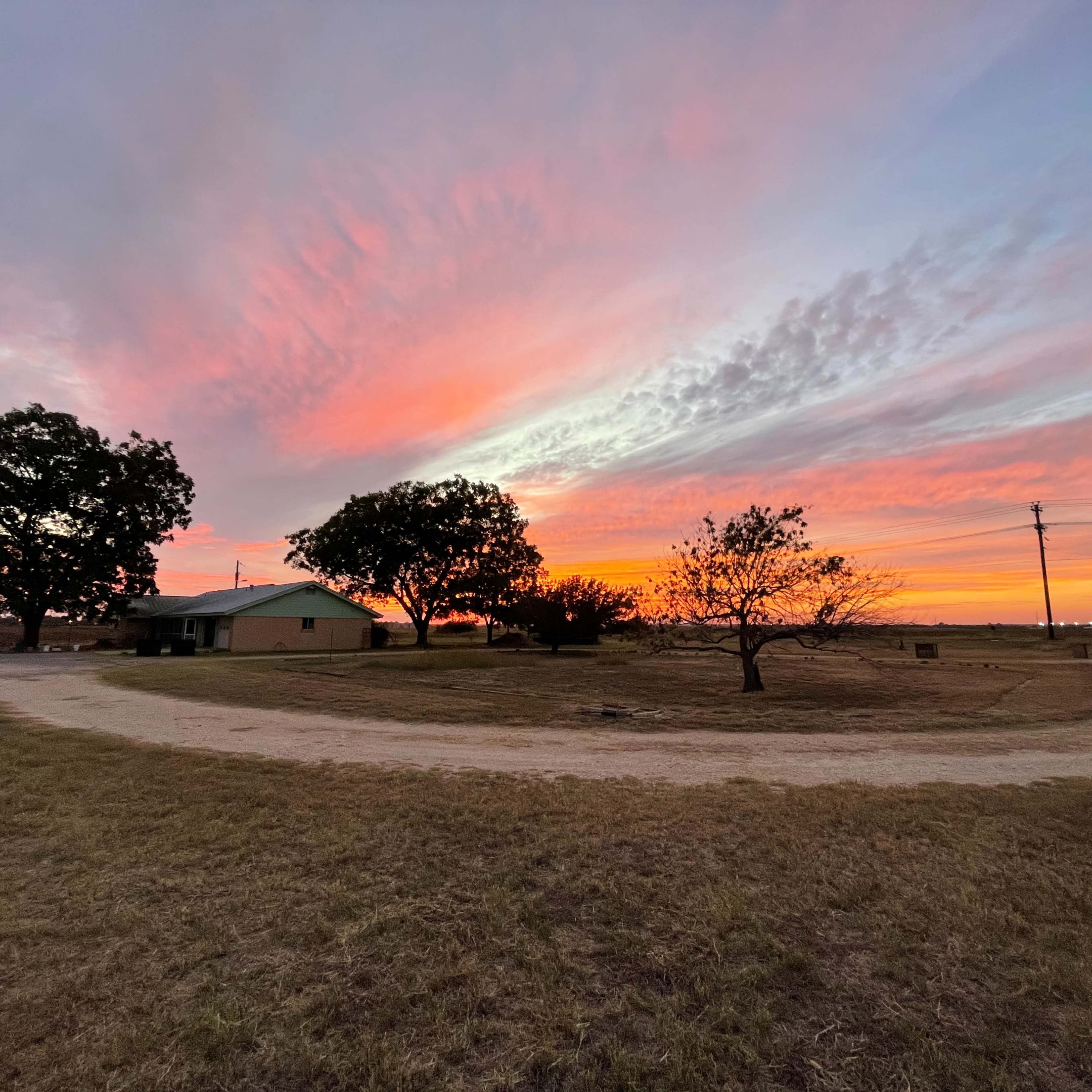 A winding dirt road leads to a house under a colorful sunset, with trees silhouetted against the sky.