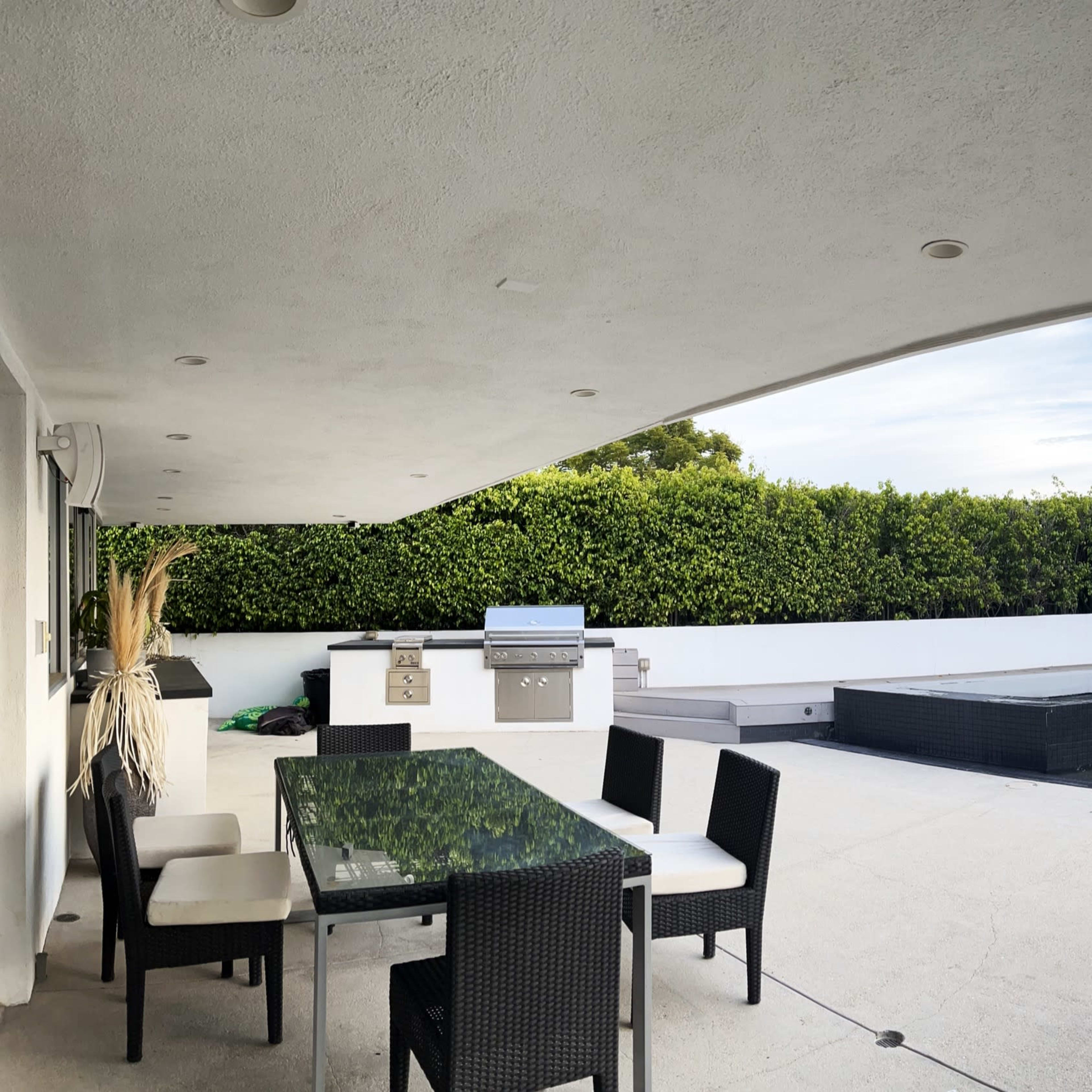 A panoramic view captures a modern outdoor space featuring a swimming pool, a barbecue area, and lounge chairs set against a backdrop of greenery and distant hills.