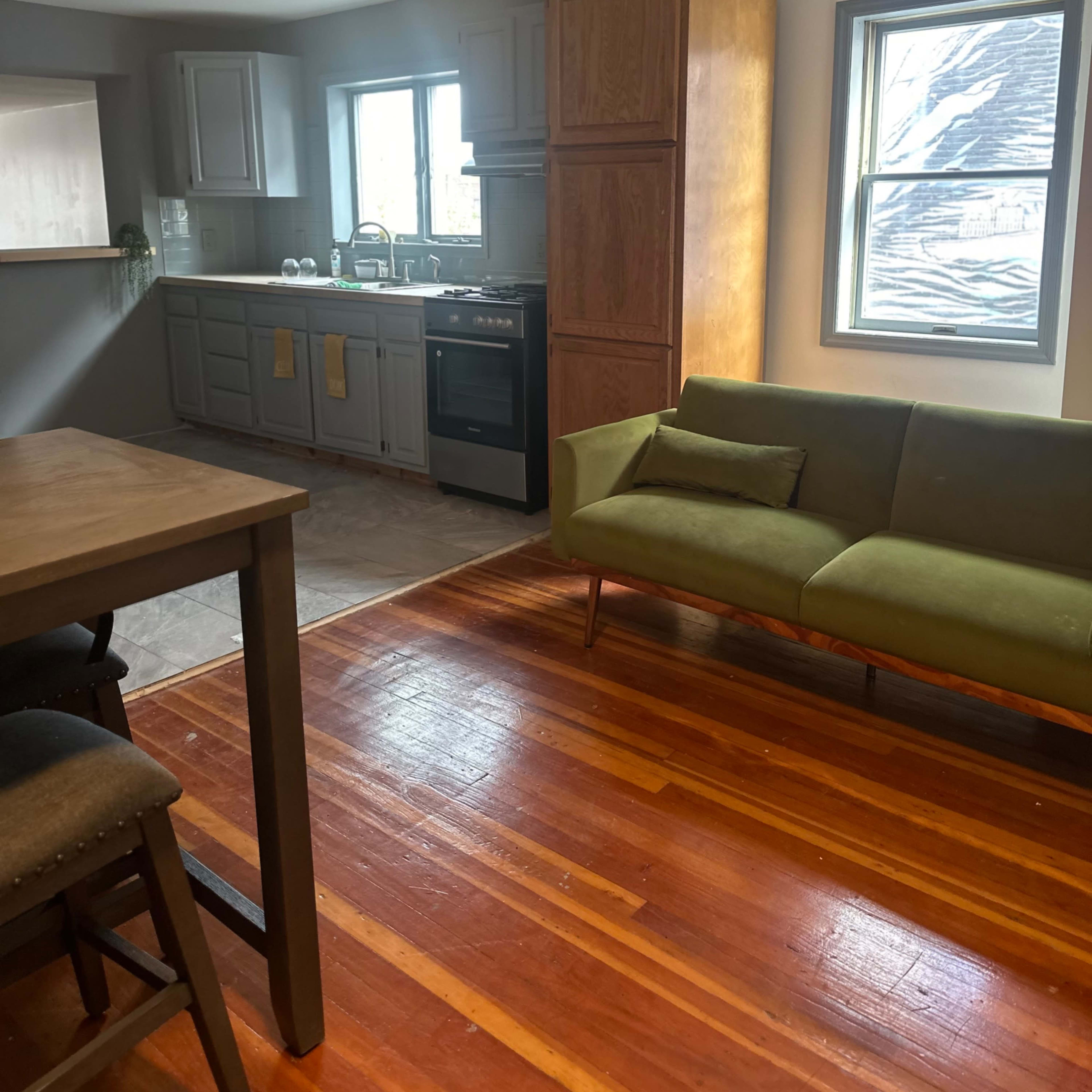 A kitchen and living area featuring wooden floors, a green couch, and cabinetry with a stove and sink.