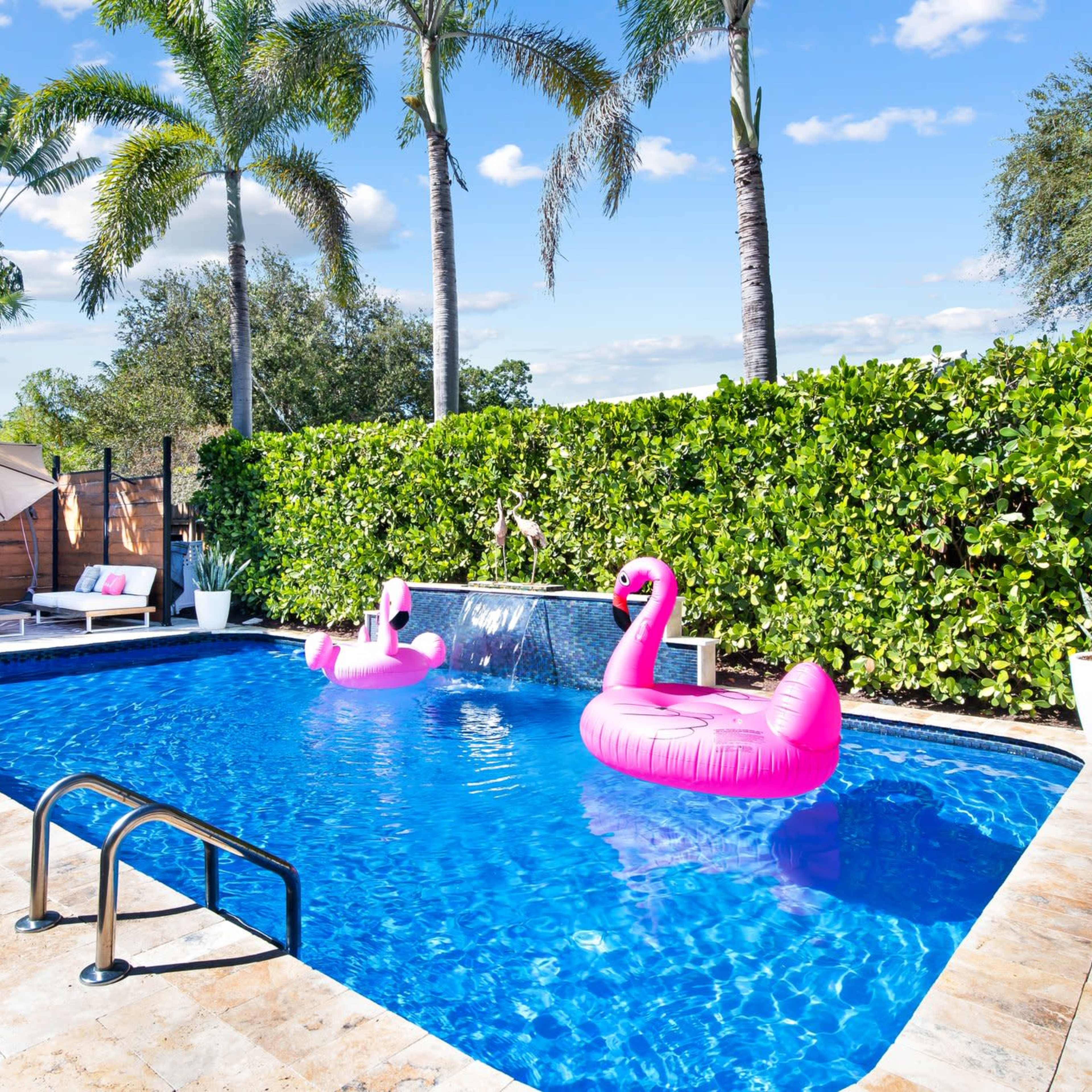 A backyard swimming pool surrounded by green hedges, two pink flamingo floaties, lounge chairs, and a shaded area with an umbrella.