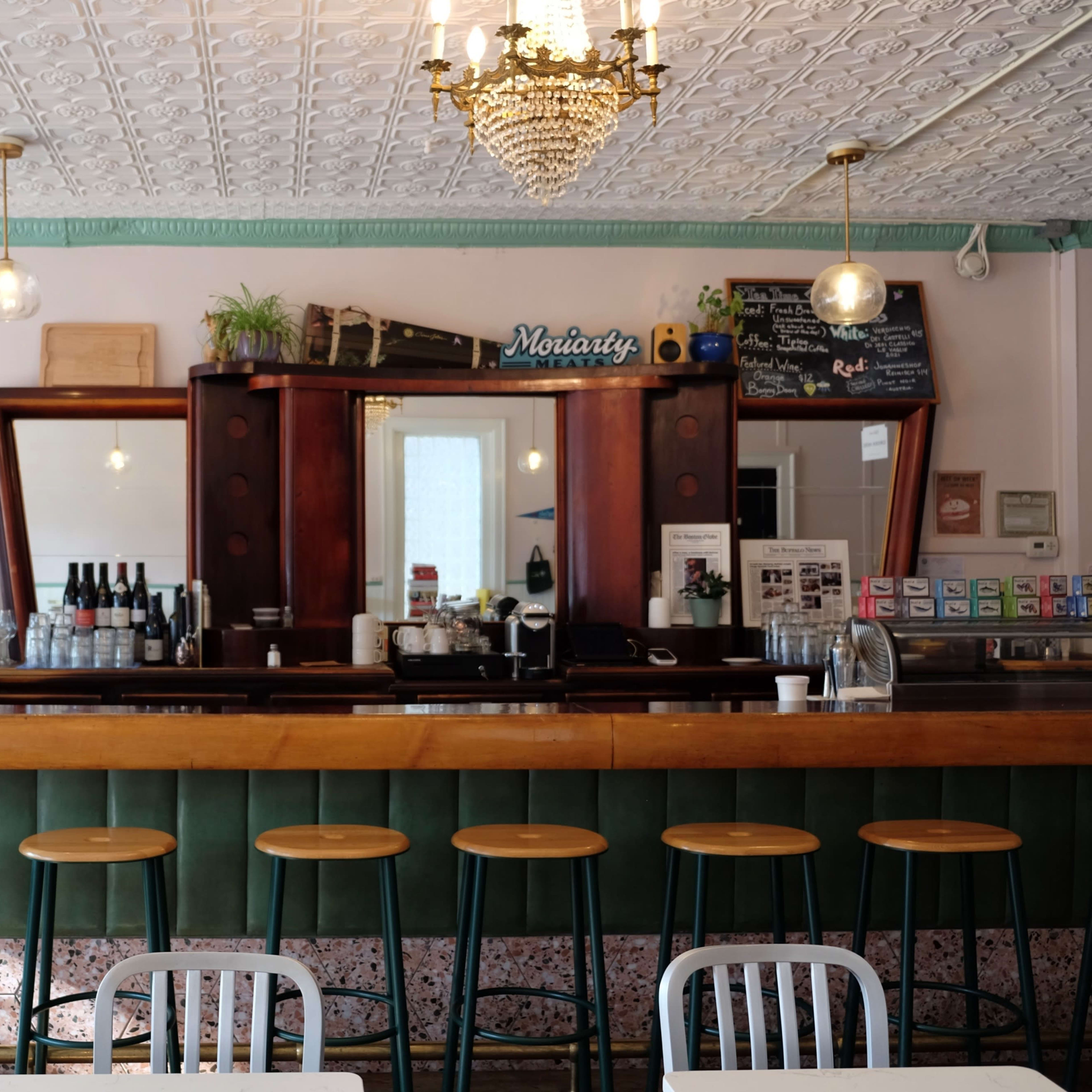 A vintage-style bar with a wooden counter, green leather seating, and a large mirror, adorned by plants and light fixtures.