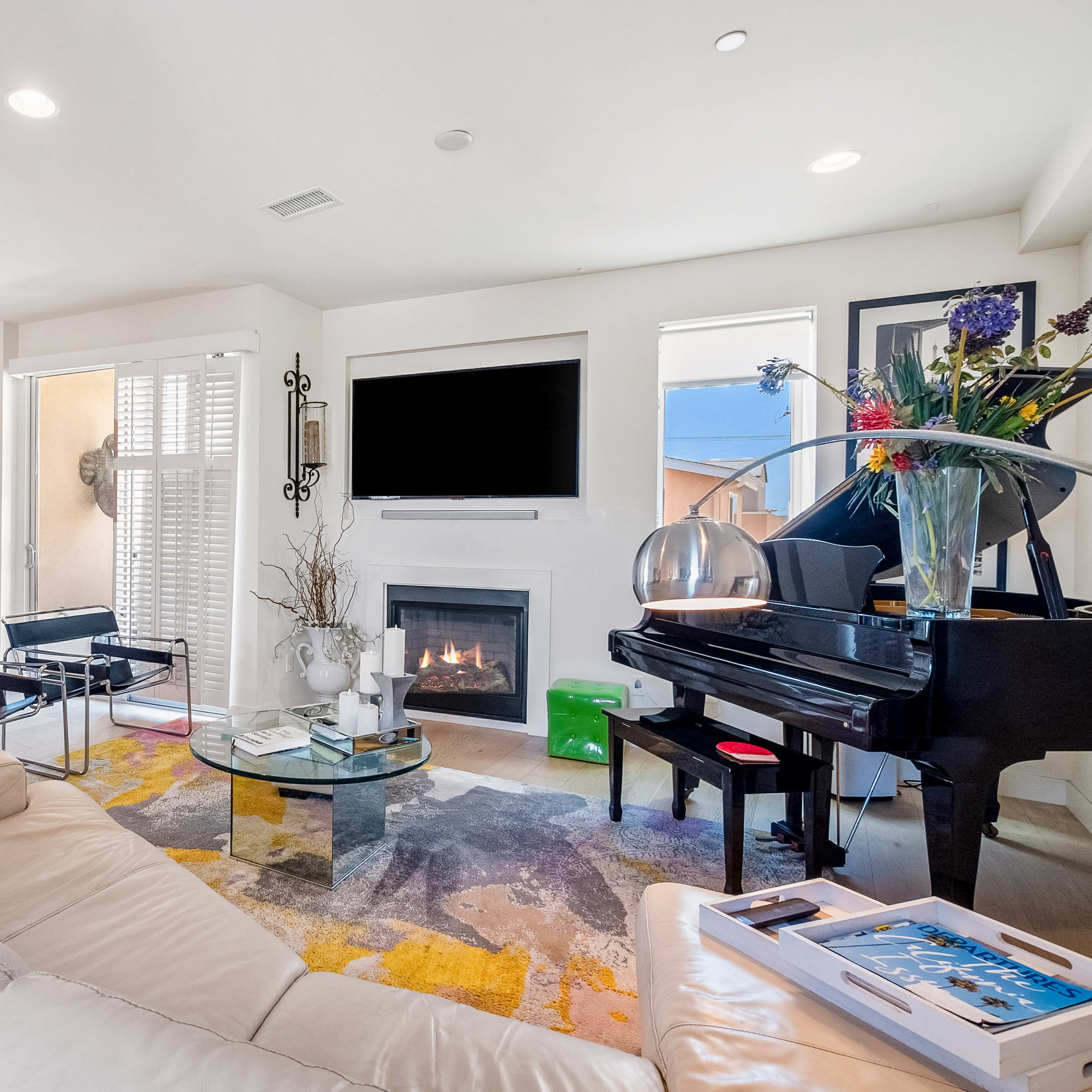 A modern living room features a white leather sofa, a grand piano, a glass coffee table, and a fireplace, with light streaming in through large windows.