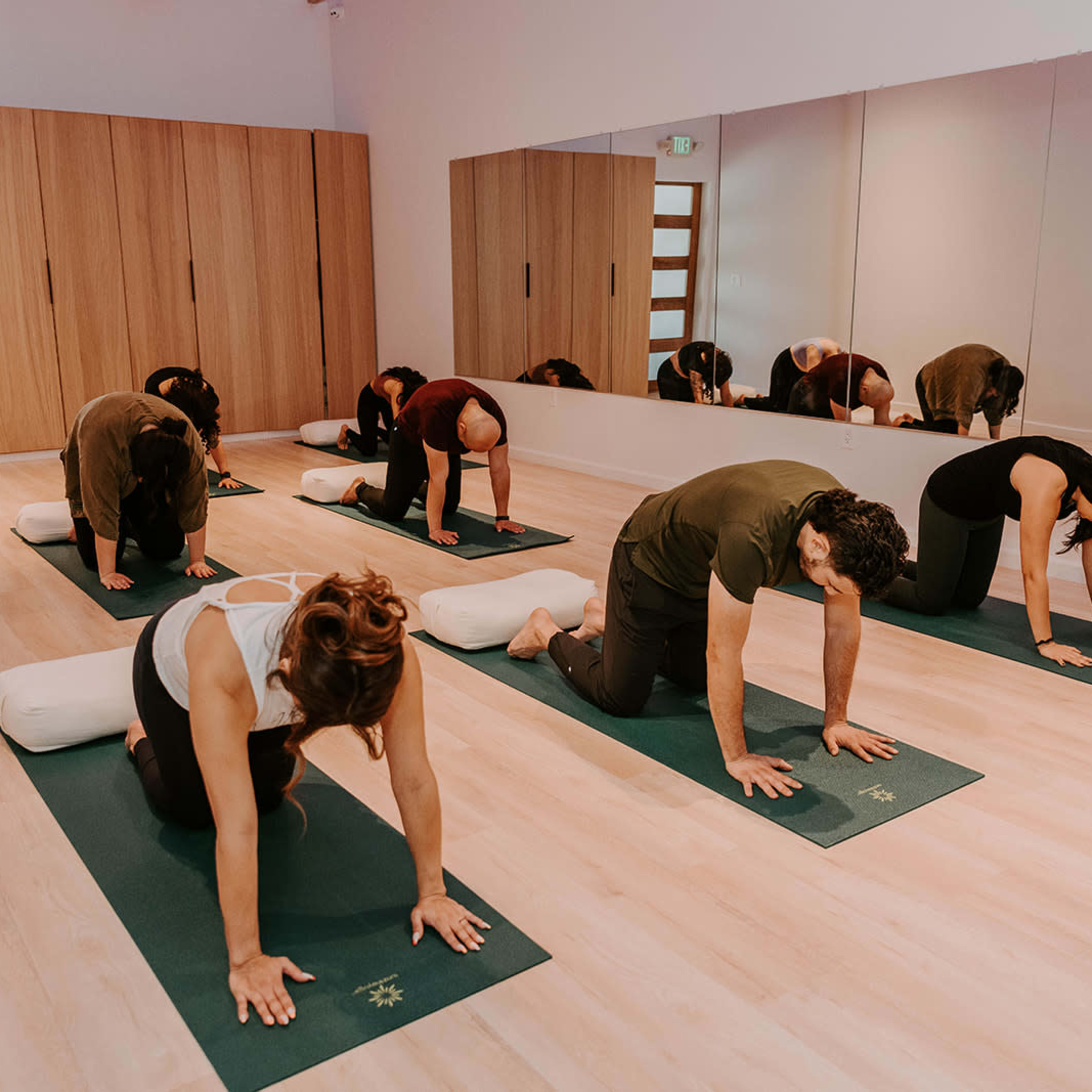 A group of people is practicing yoga on mats in a well-lit studio with mirrors and wooden paneling.