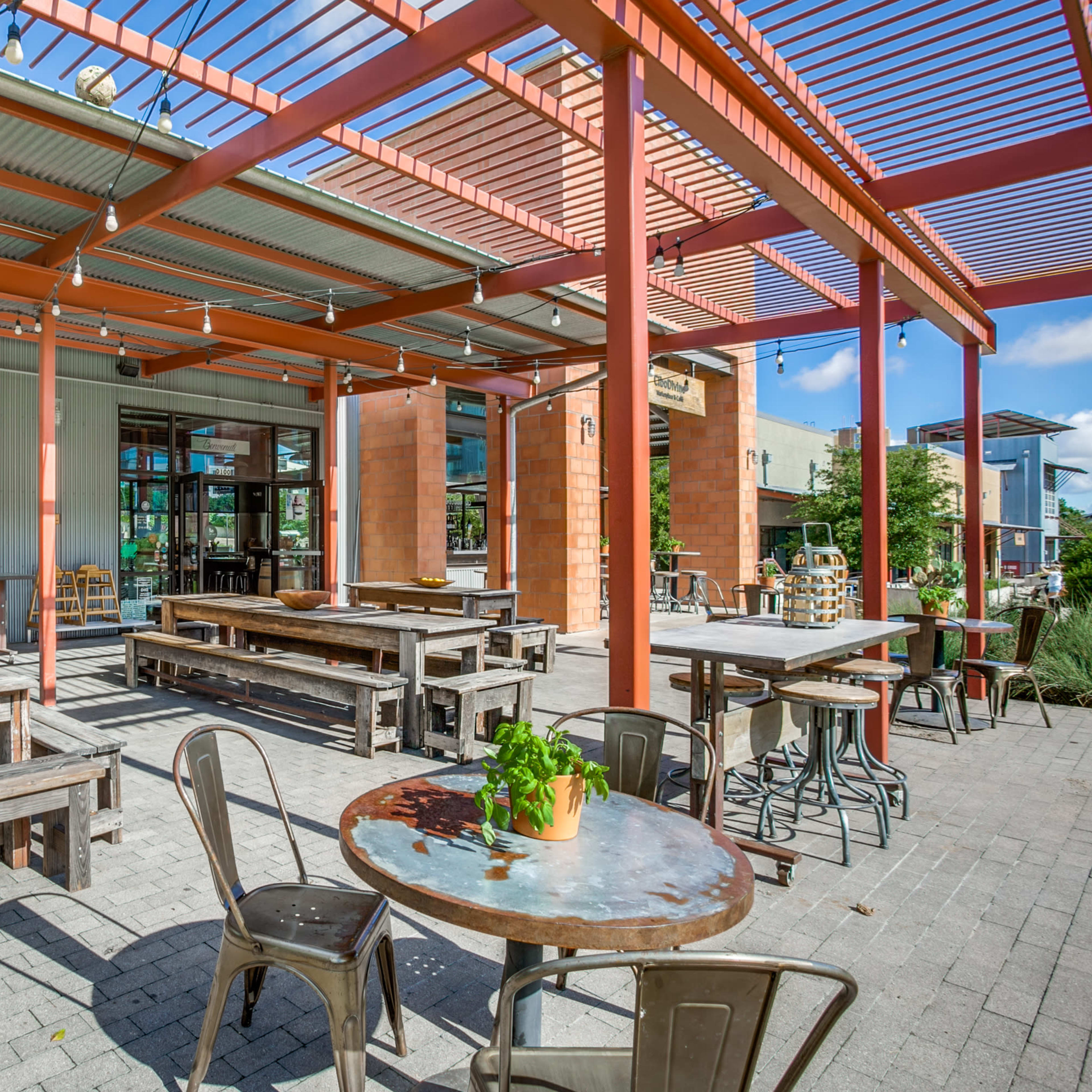 An exterior dining area with wooden tables and metal chairs under a red framed awning.