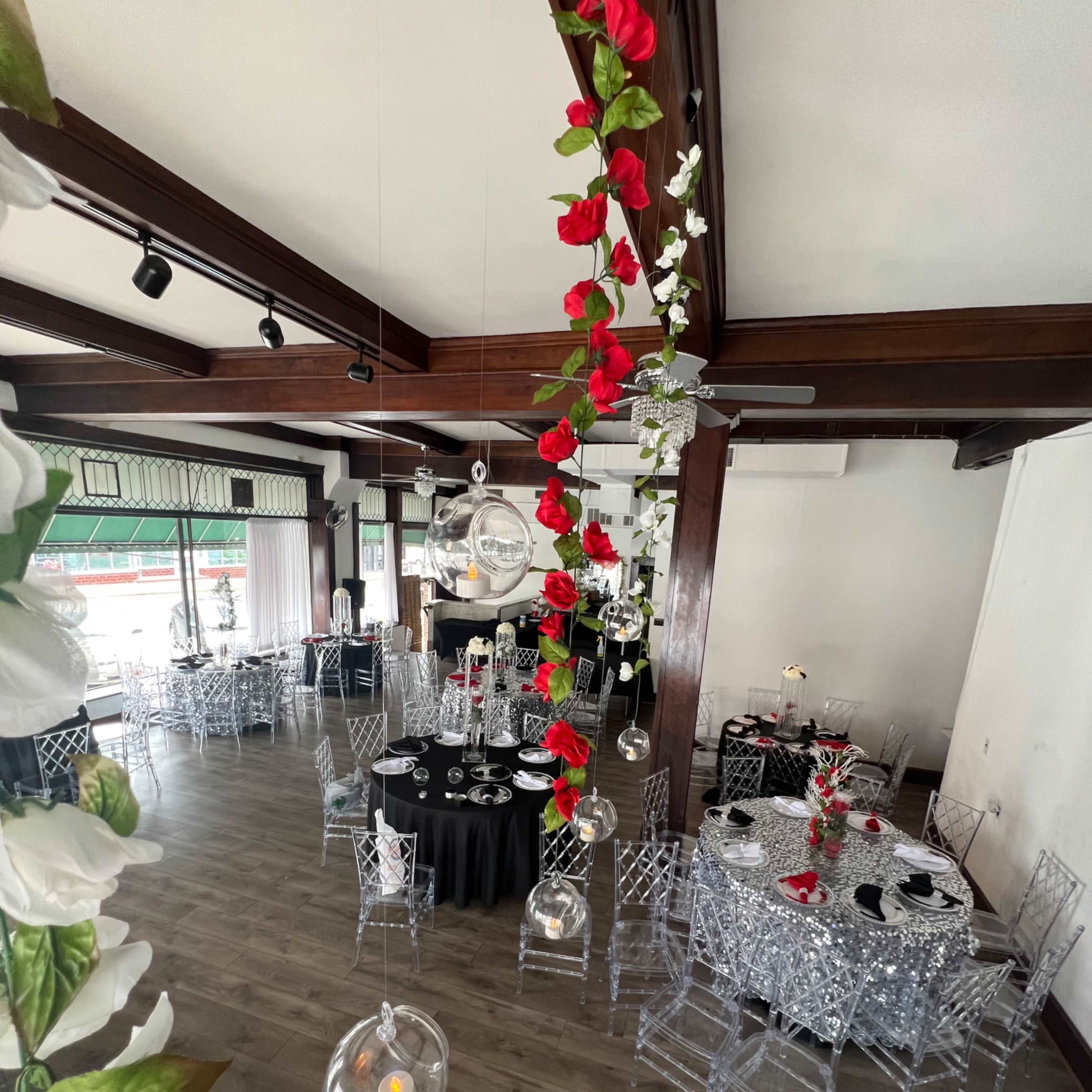 A decorated banquet hall with tables elegantly set for an event, featuring black and silver tablecloths, transparent chairs, and hanging floral arrangements.