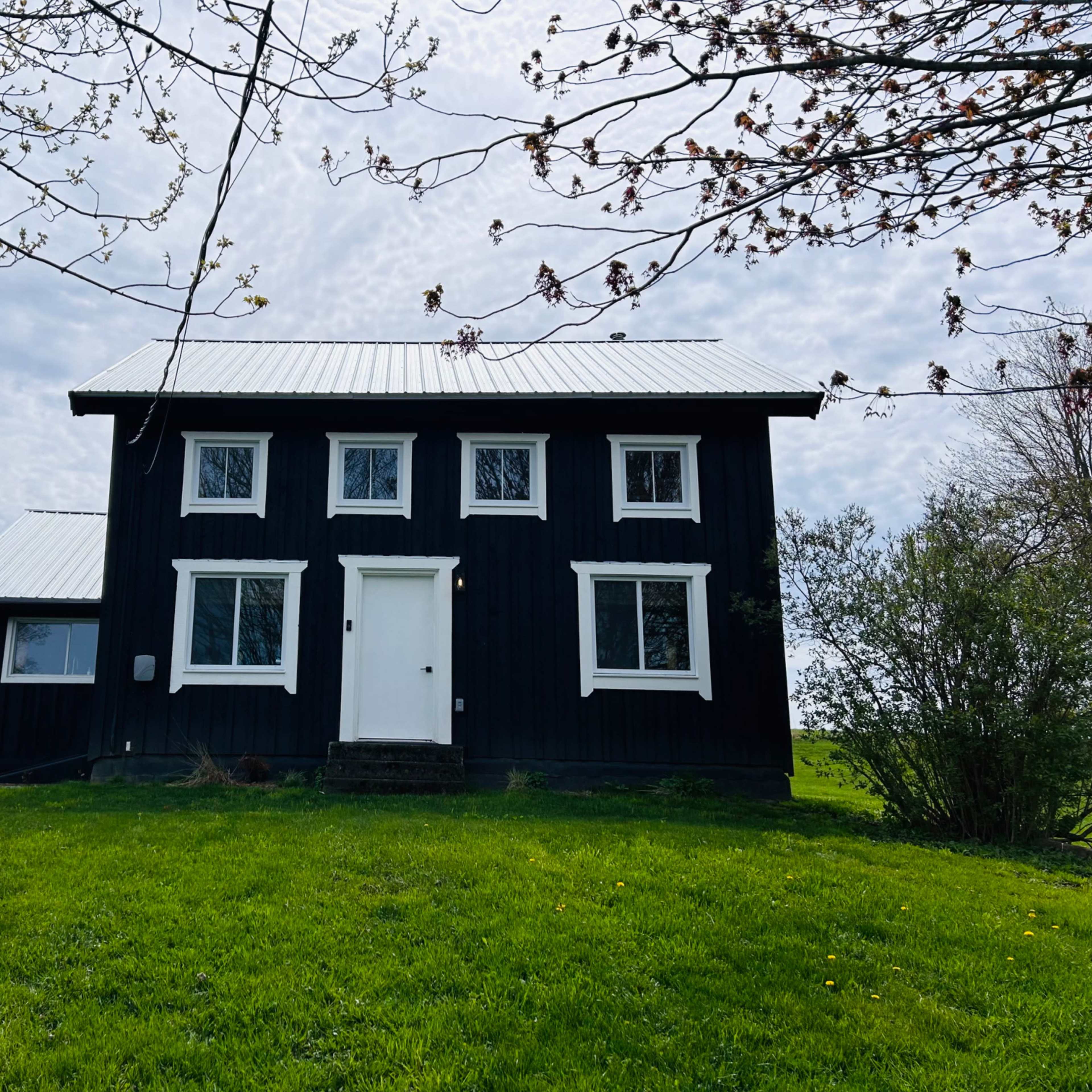 A black two-story house with a white roof and several windows sits on a green lawn under a cloudy sky.