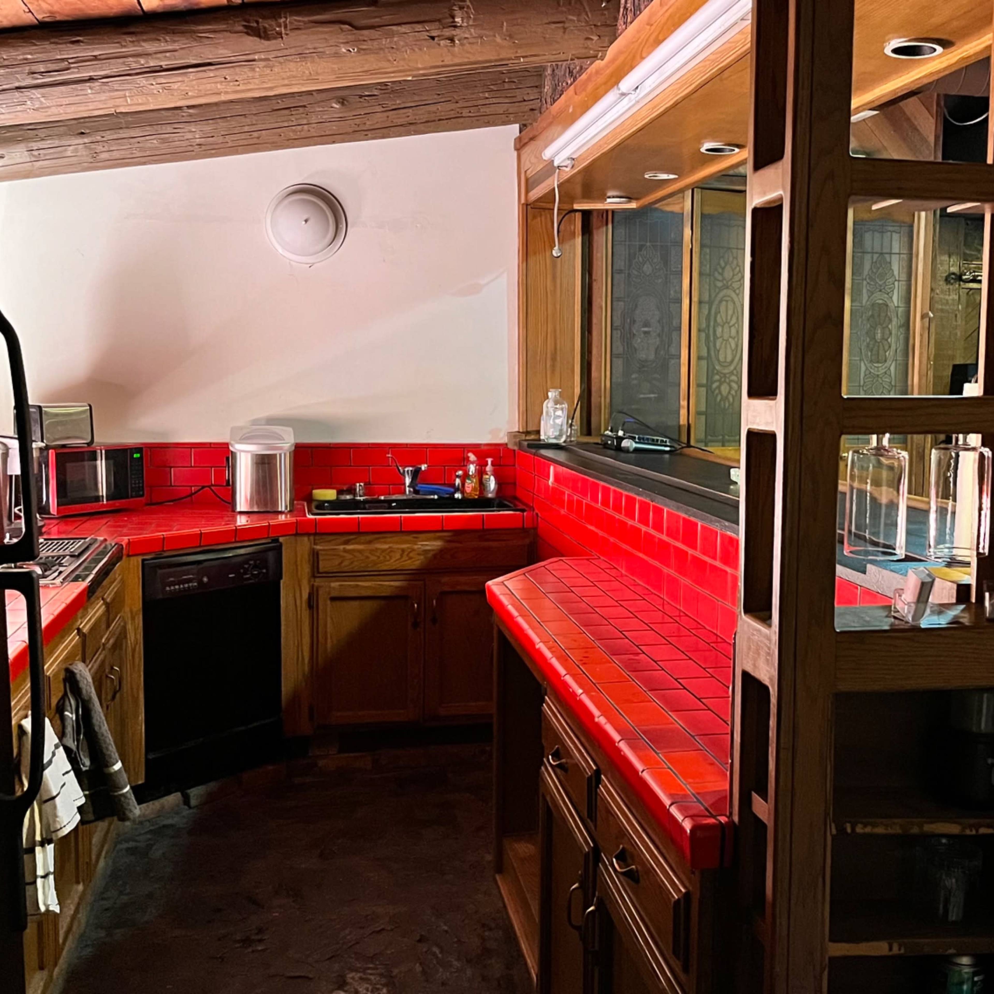 A kitchen with wooden cabinets, a red tiled countertop, and an open layout leading to a room with brick walls and artwork.