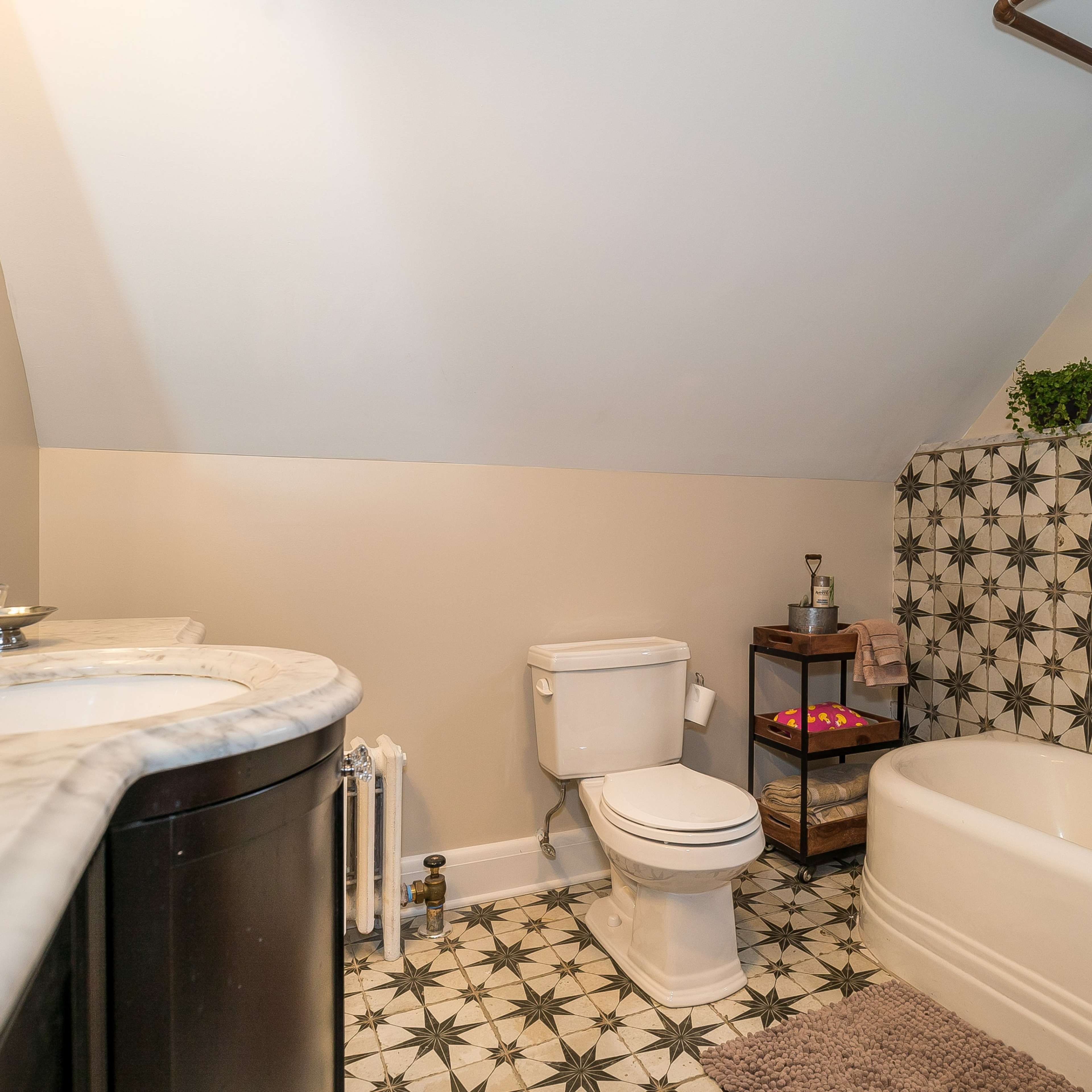 The bathroom features a marble-topped vanity, a toilet, a clawfoot bathtub, and patterned tile flooring underneath a sloped ceiling.