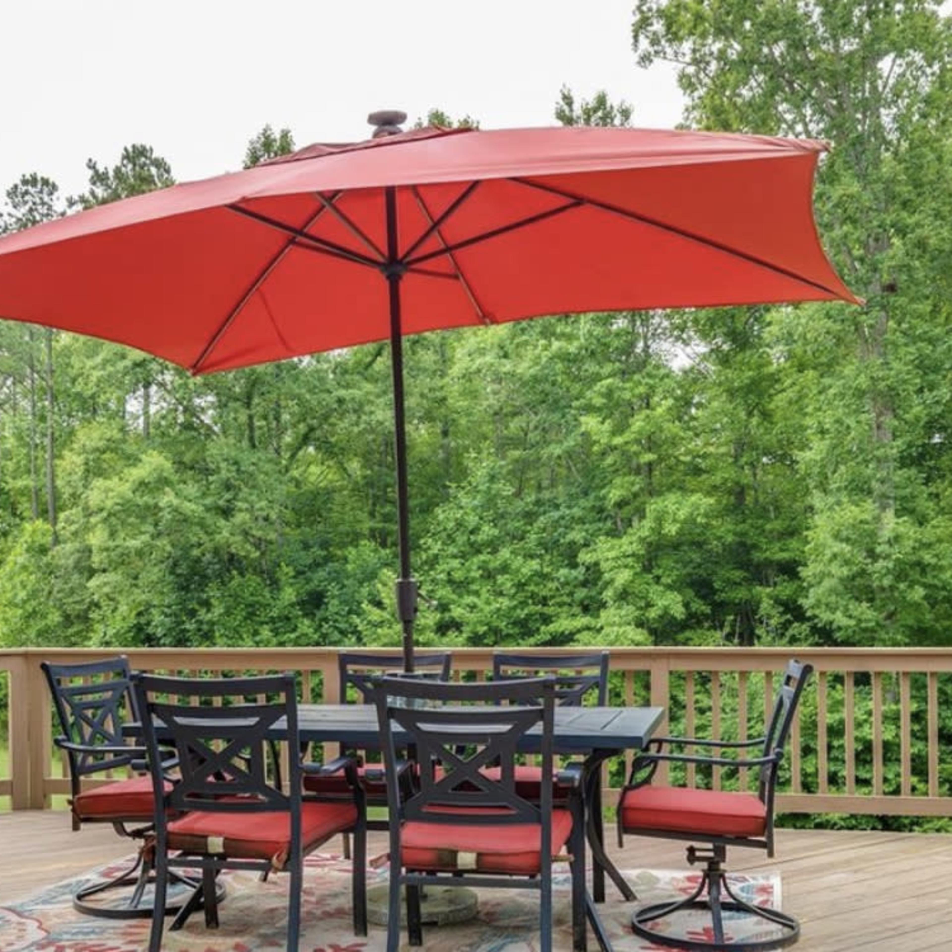 A red umbrella shades a round table surrounded by chairs on a wooden deck overlooking a lush green landscape.