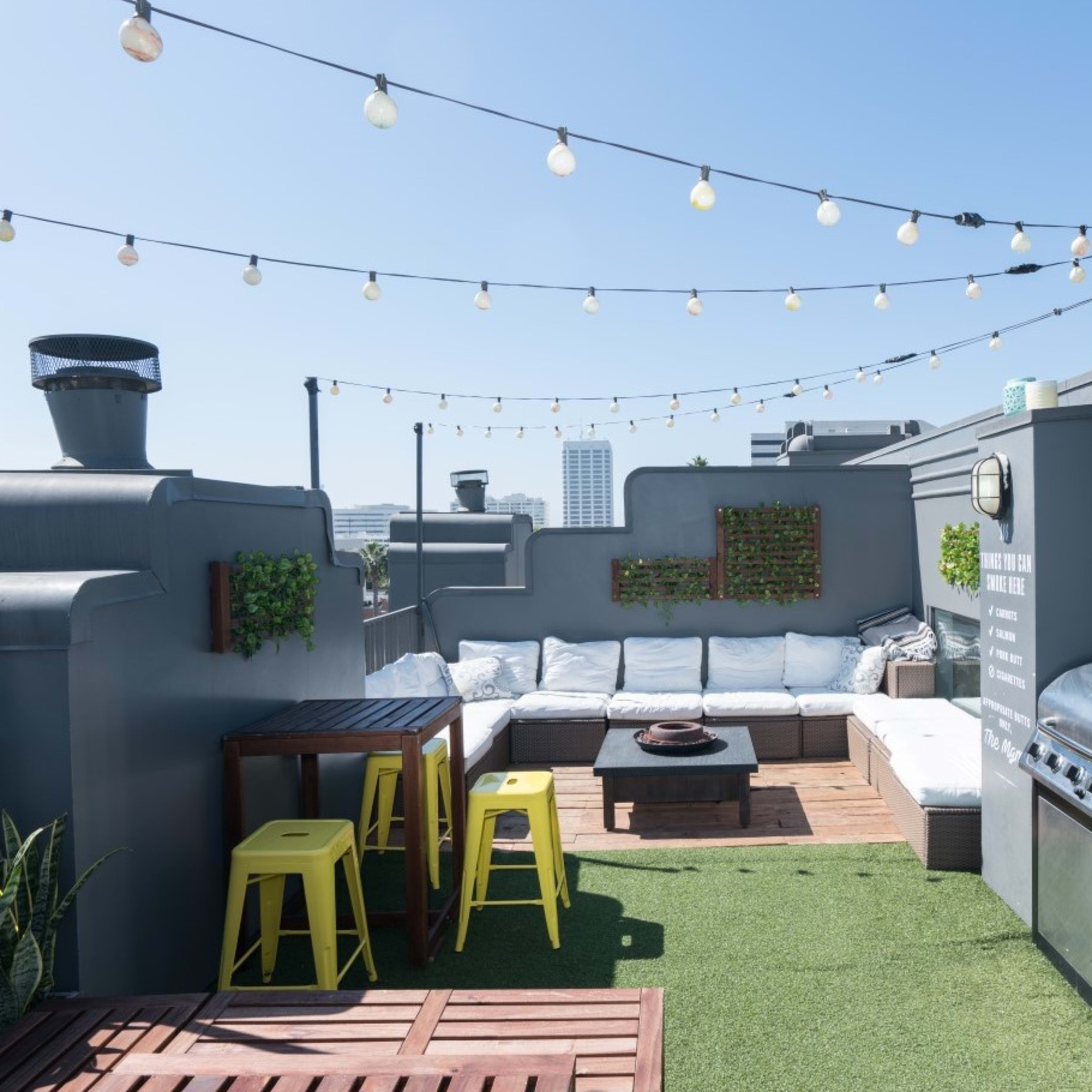 A rooftop patio with seating, a barbecue grill, string lights, and green plants against a clear blue sky.
