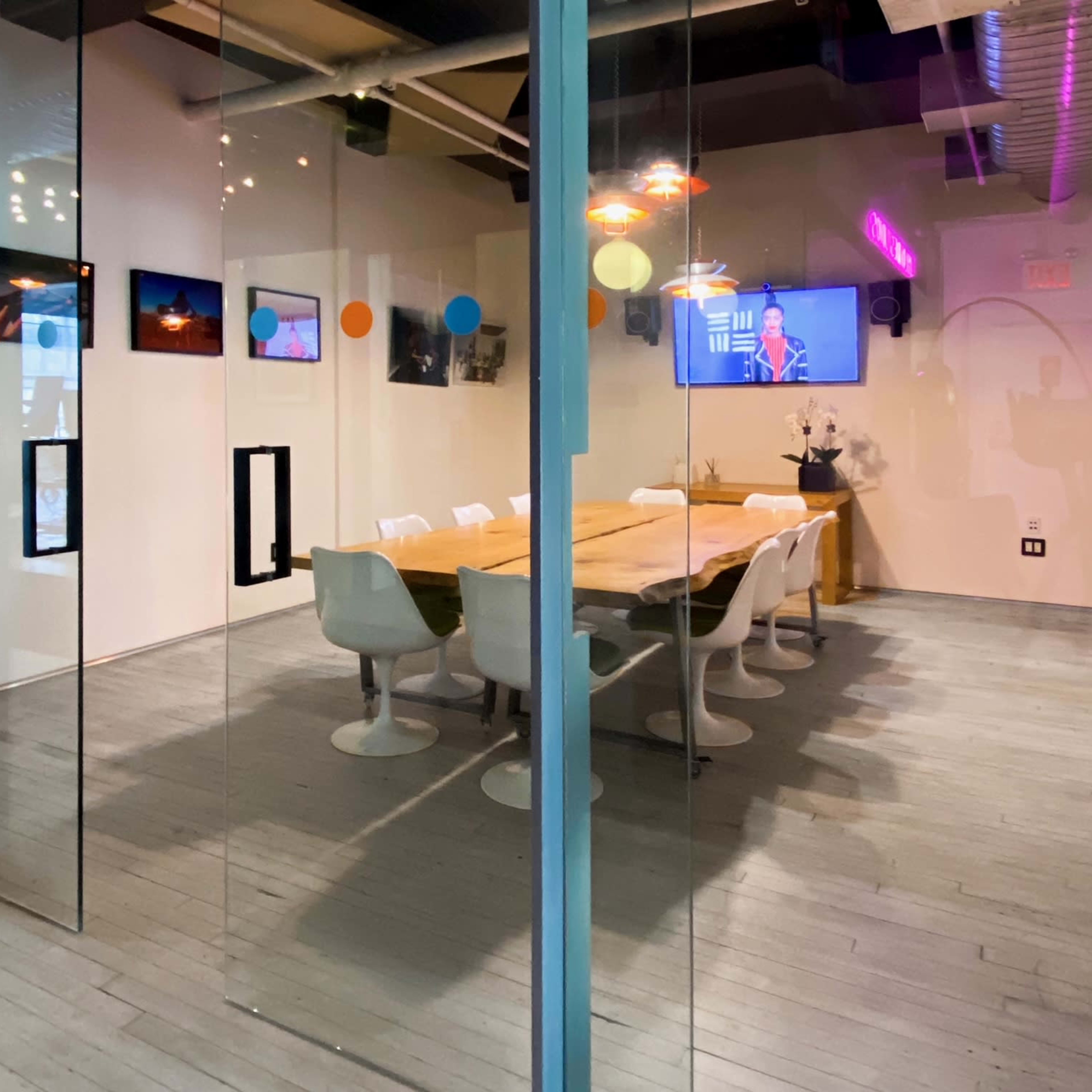 A conference room with a large wooden table and white chairs is visible through glass walls, featuring artworks on the walls and a television displaying a colorful screen.