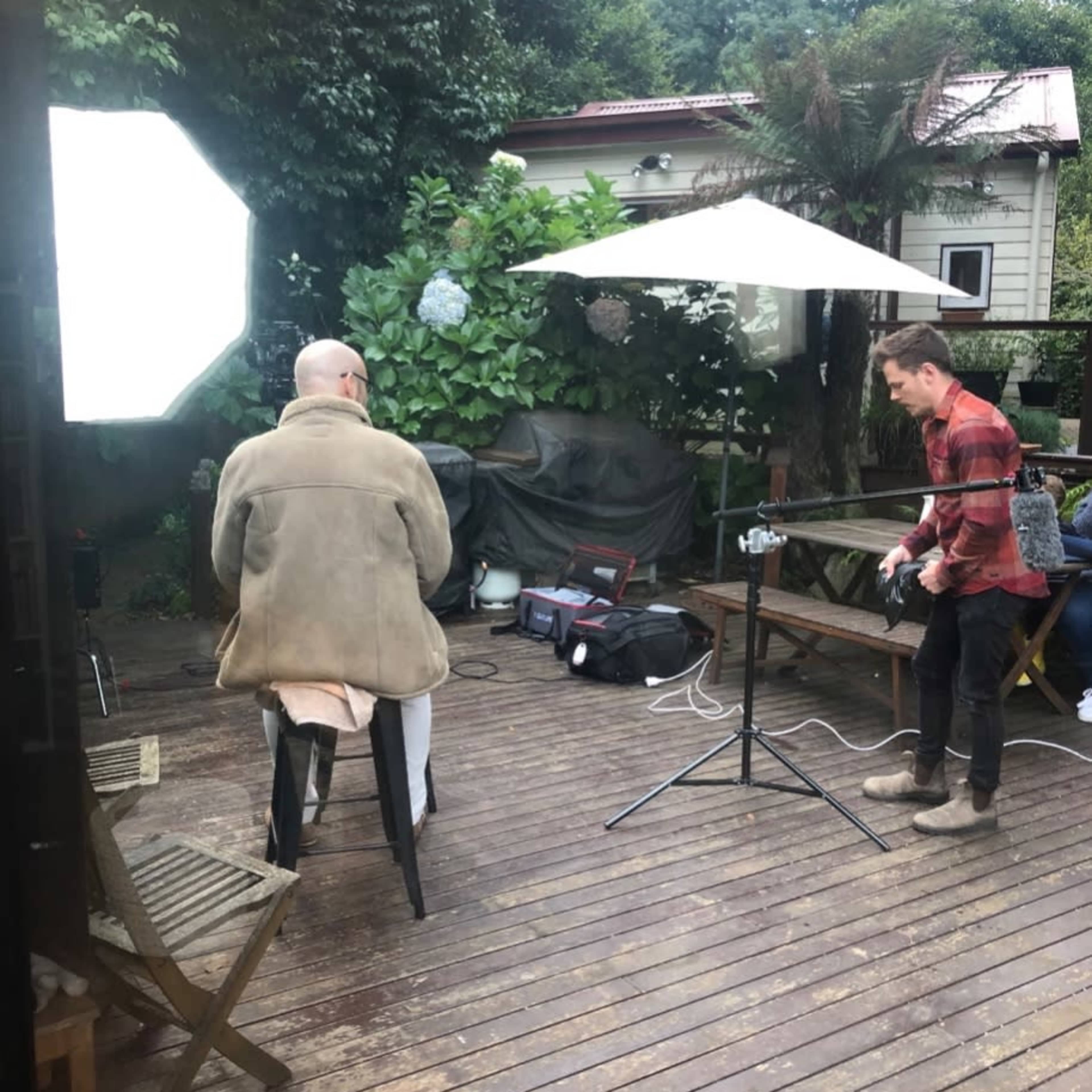 A person sits on a stool in a backyard while another person adjusts a camera setup under an umbrella.