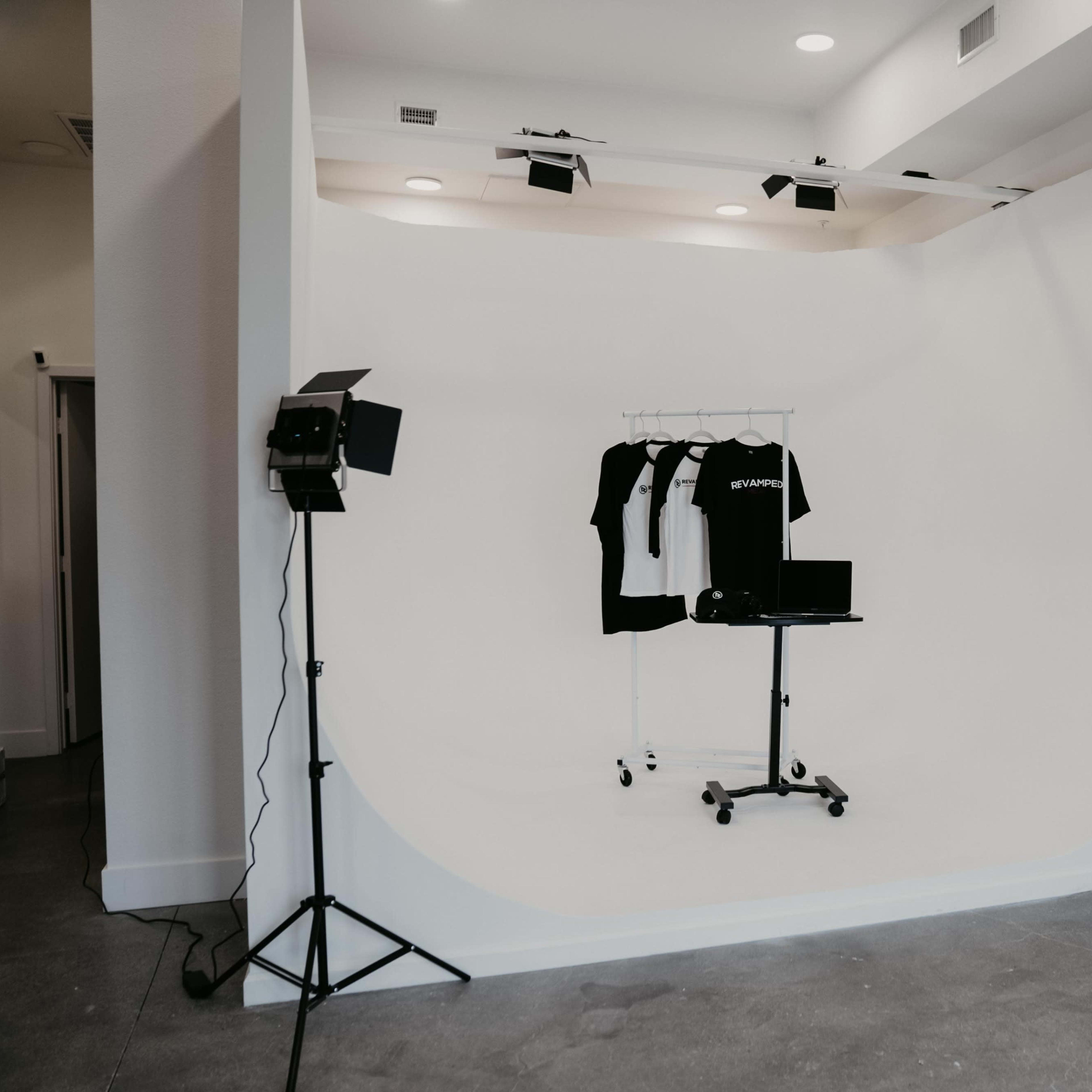 A minimalist photography studio with a white backdrop, a clothing rack holding black and white shirts, and lighting equipment arranged on either side.