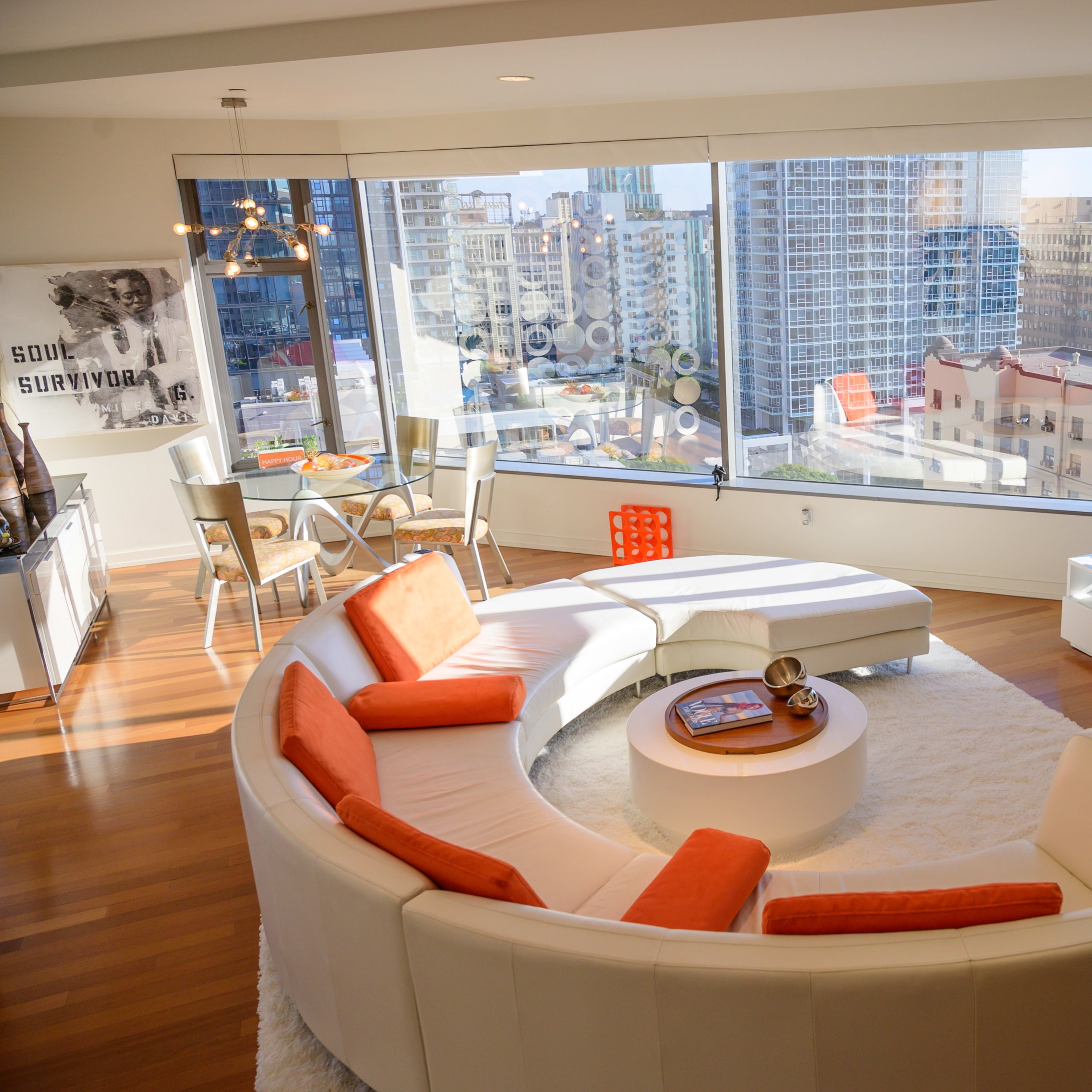 A modern living room with a circular white sofa, orange accents, a television, and a large window offering a city view.