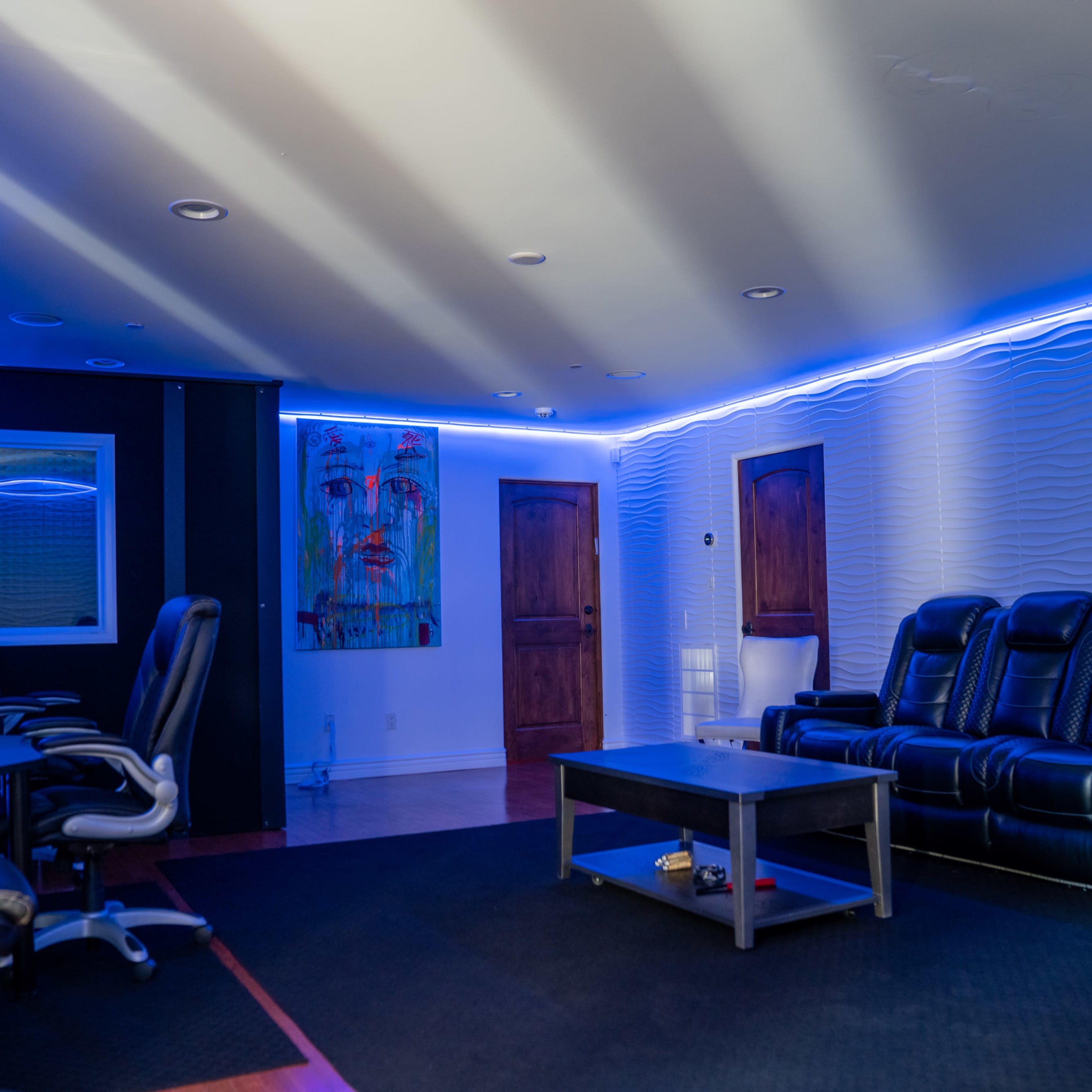A dimly lit room features a black sectional sofa, a coffee table, and computer chairs arranged in front of glowing blue LED lights along the walls.