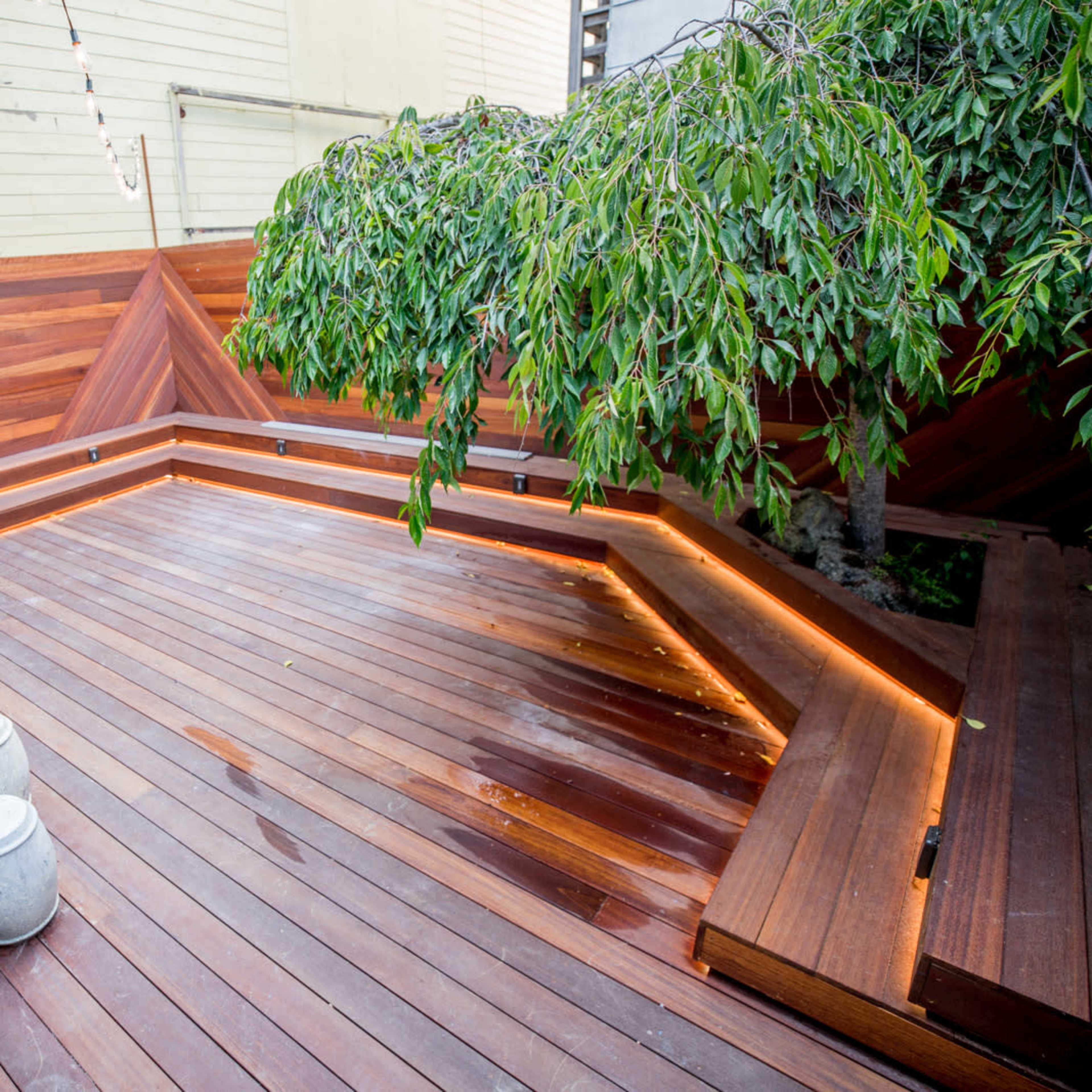 A wooden deck with built-in seating and soft ambient lighting, featuring a small table and a tree in the corner.