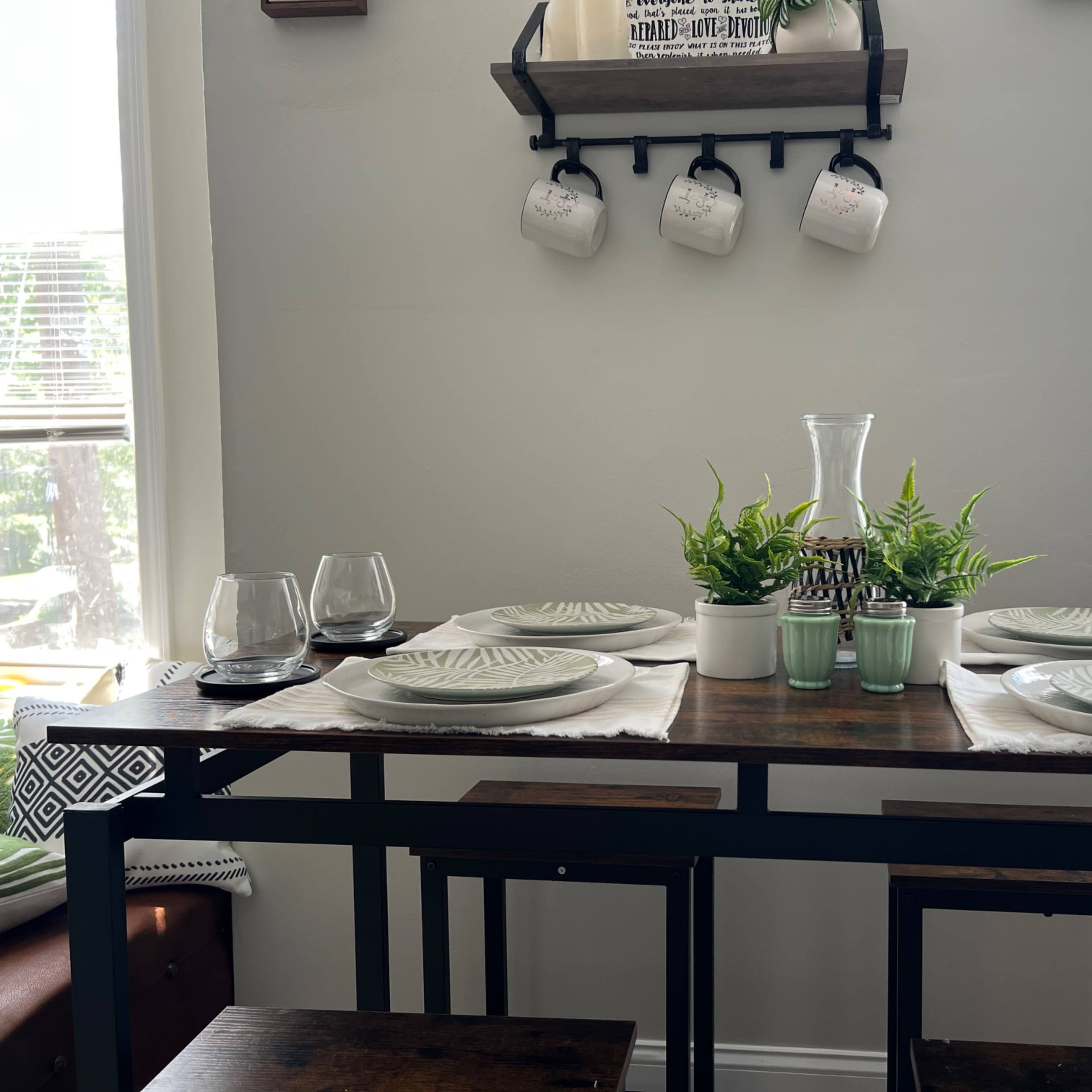 A dining table is set with plates, glasses, and decorative plants, positioned near a wall with shelves displaying mugs.