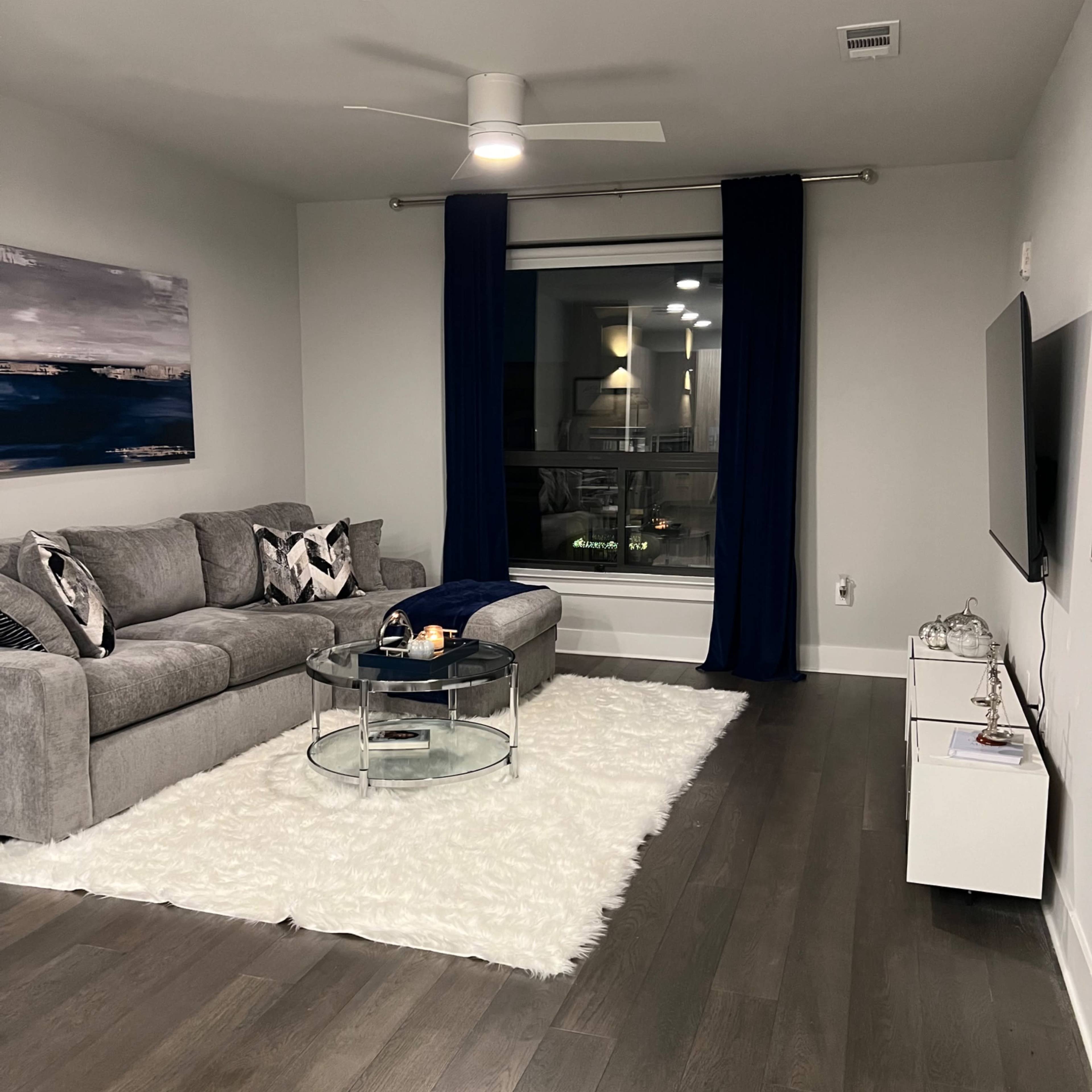 A modern living room with a gray sectional sofa, a glass coffee table on a white rug, and large windows dressed with dark blue curtains.