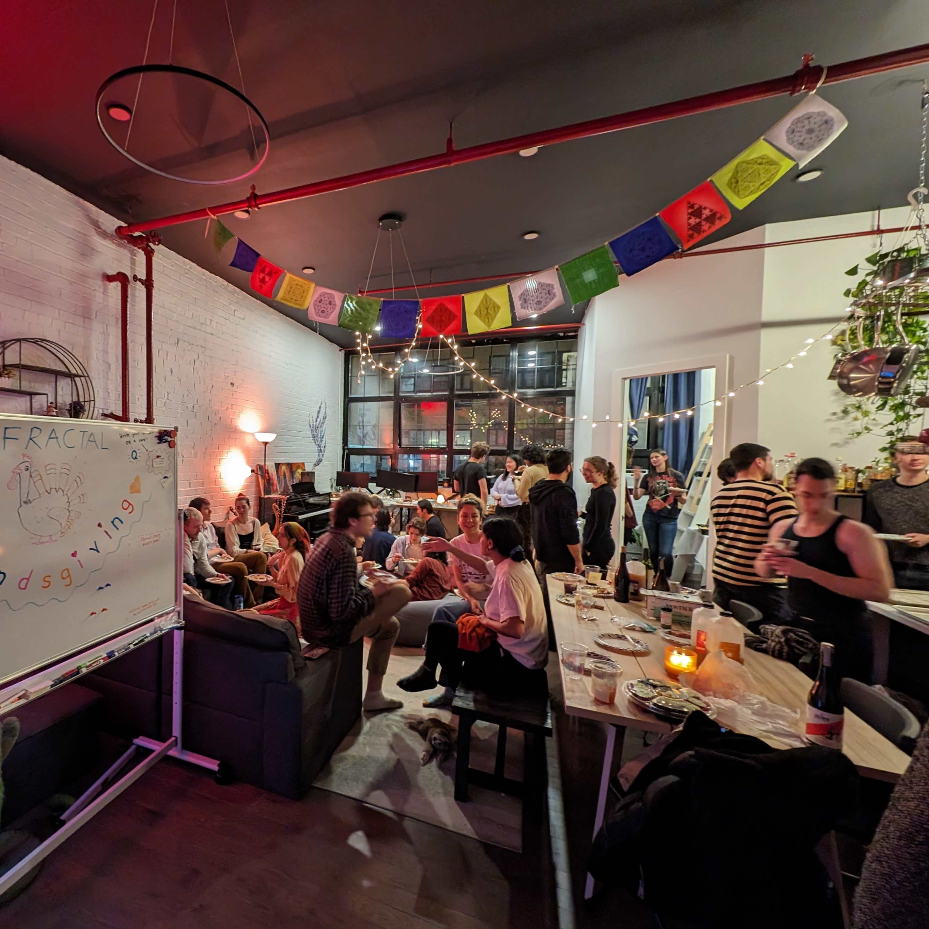 A lively gathering is taking place in a room decorated with colorful prayer flags, where people are seated at tables and conversing.