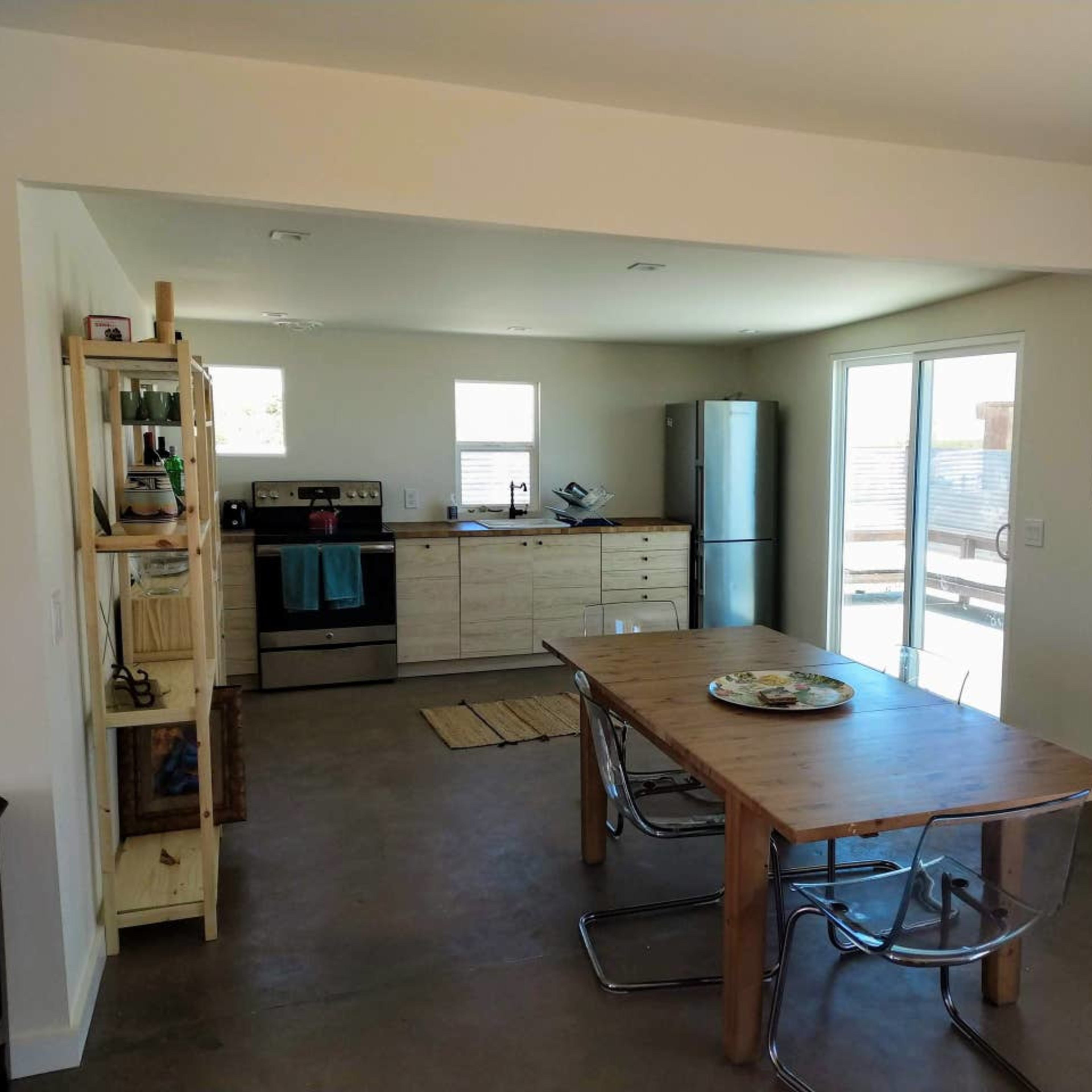A minimalist kitchen and dining area with a wooden table, stainless steel appliances, and a sliding glass door leading outside.