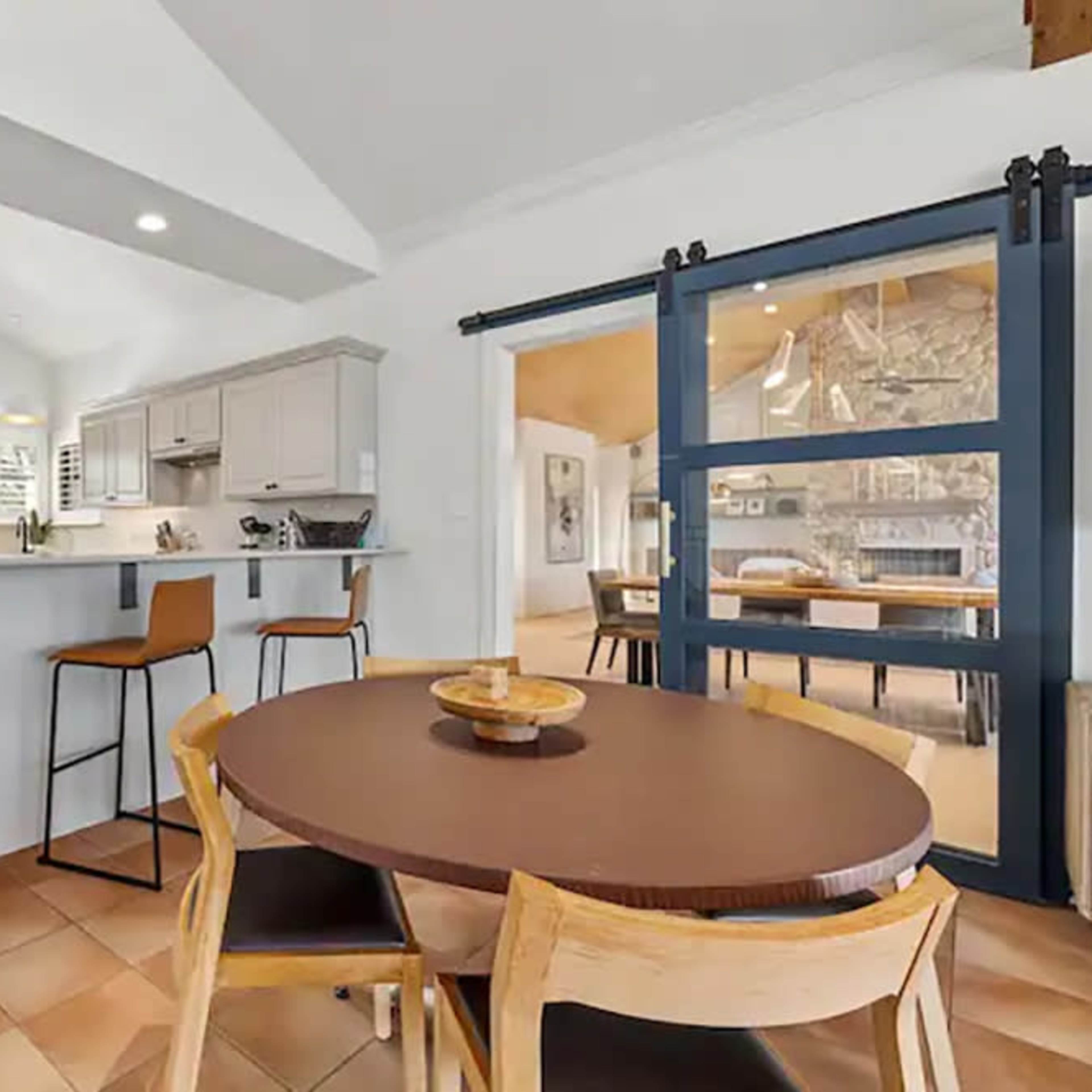 A modern kitchen and dining area with a round table and bar stools, accompanied by light-colored cabinets and tiled flooring.