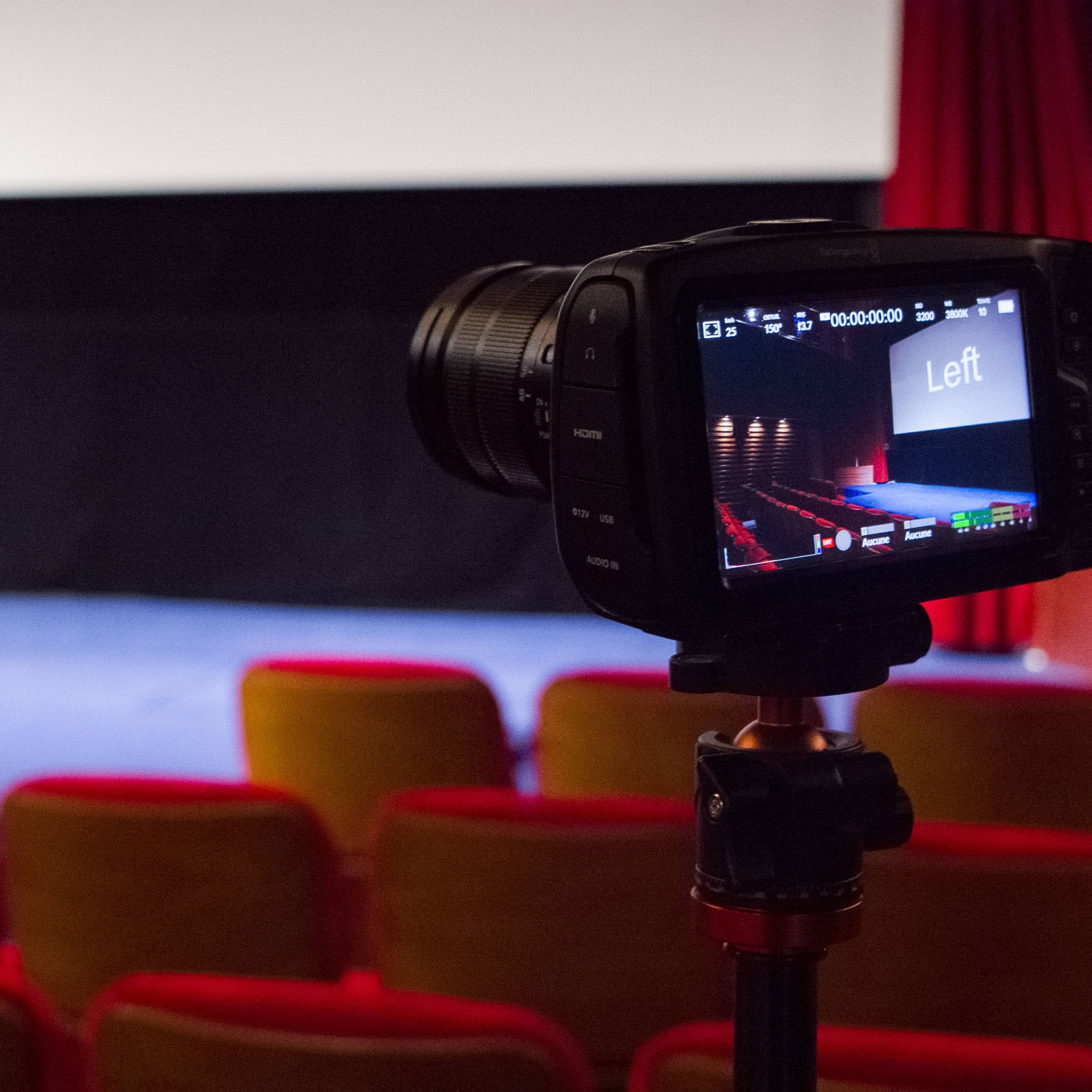 A camera is positioned on a tripod, focused on an empty cinema screen displaying the word "Left," with red theater seats visible in the foreground.