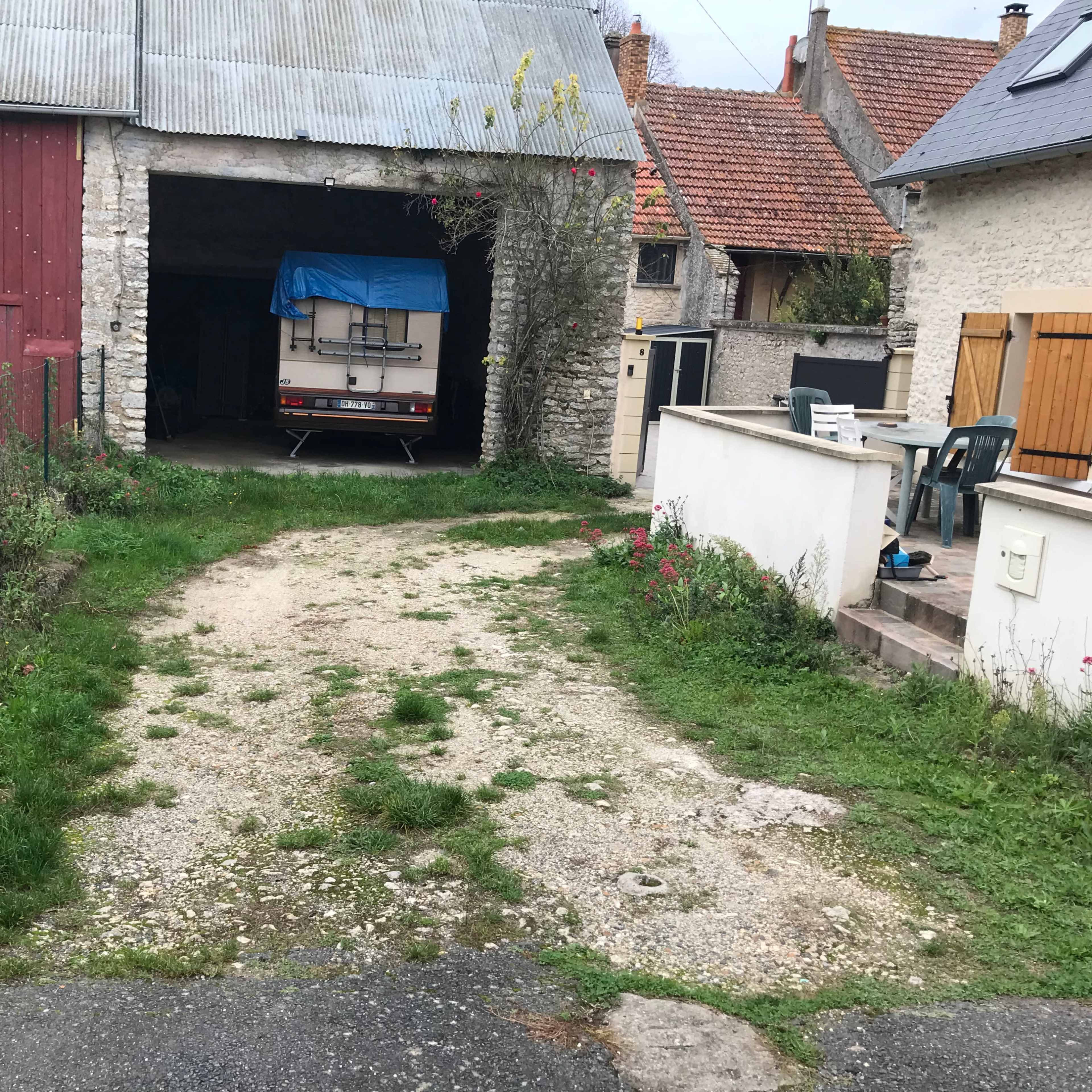 A gravel path leads to a garage with a blue tarp covering a trailer, surrounded by overgrown grass and scattered flowers.