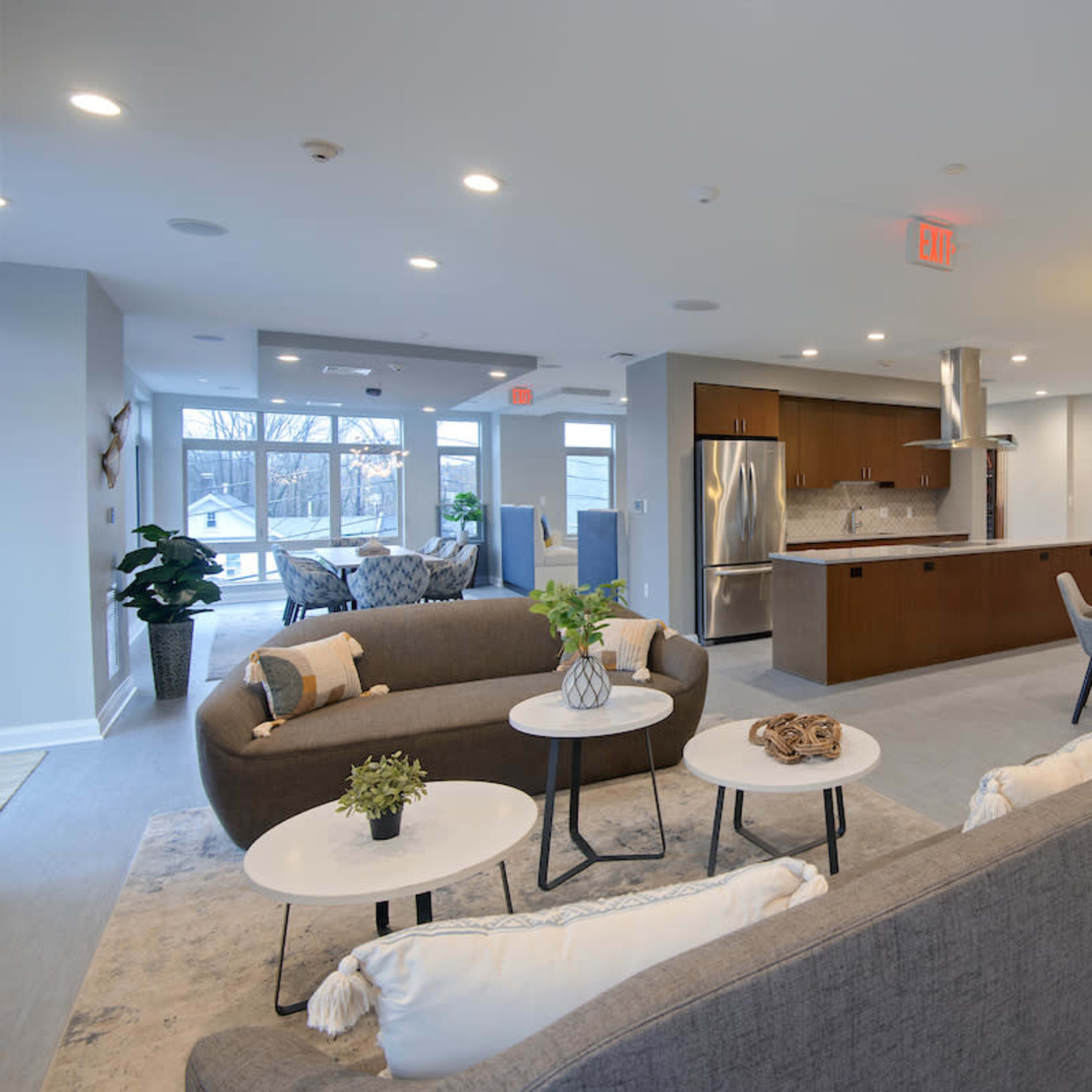 A modern communal lounge features seating areas with sofas and tables, a kitchen bar, and dining space with chairs, illuminated by pendant lights.
