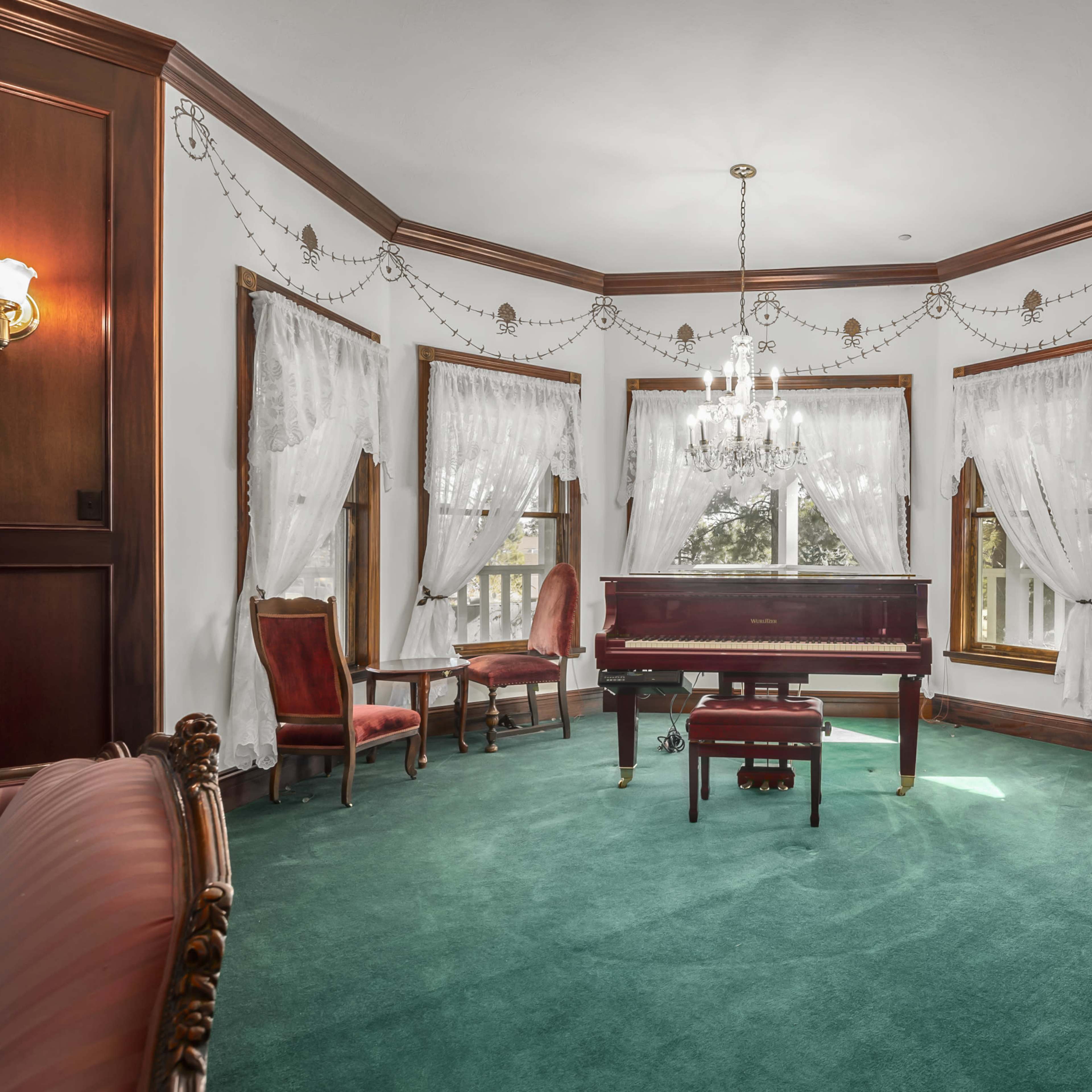The room features a grand piano centered on a green carpet, surrounded by elegant seating and large windows adorned with draped curtains.